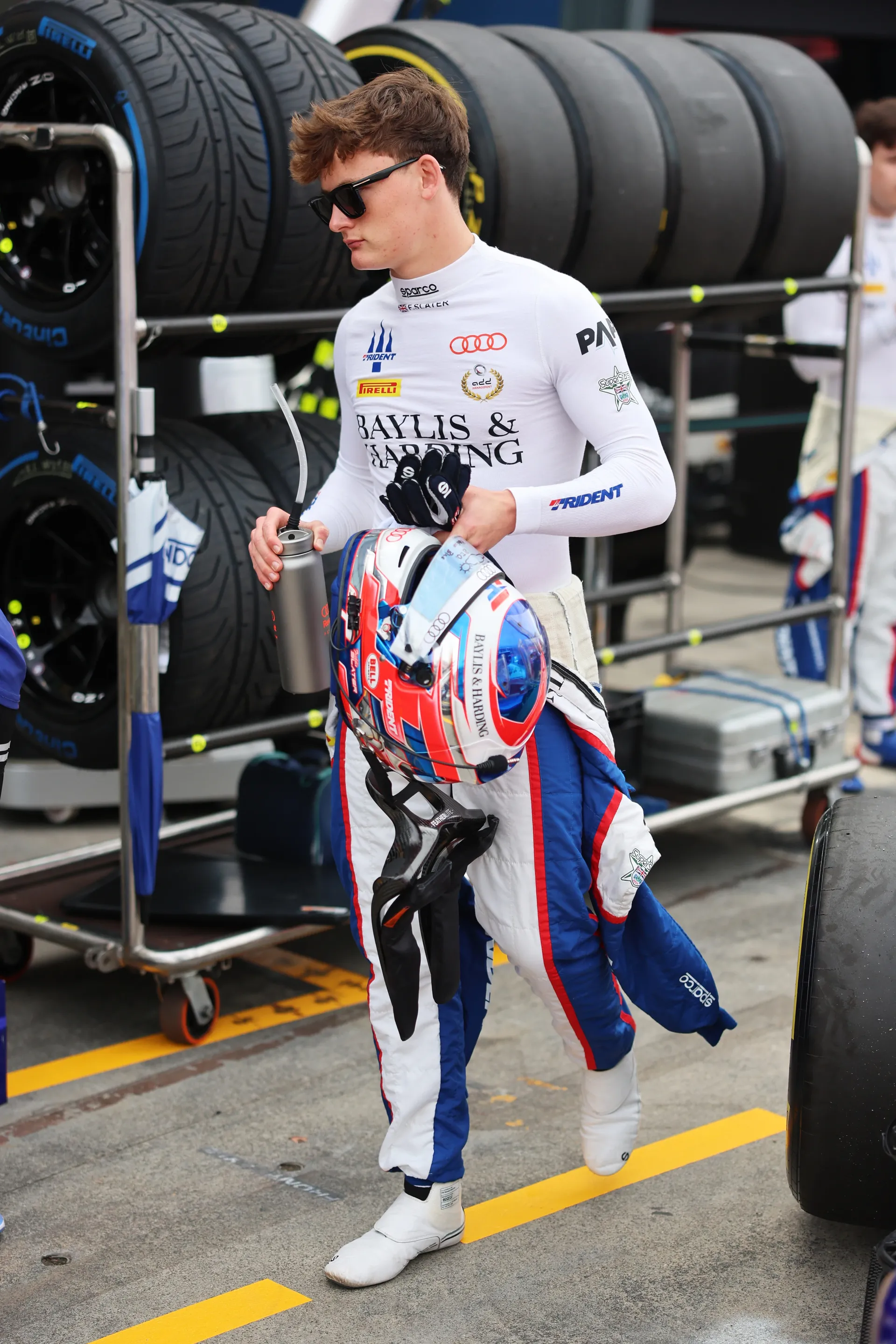 Freddie Slater carrying his helmet and water bottle walking past stacked tires in the Formula 3 pit lane
