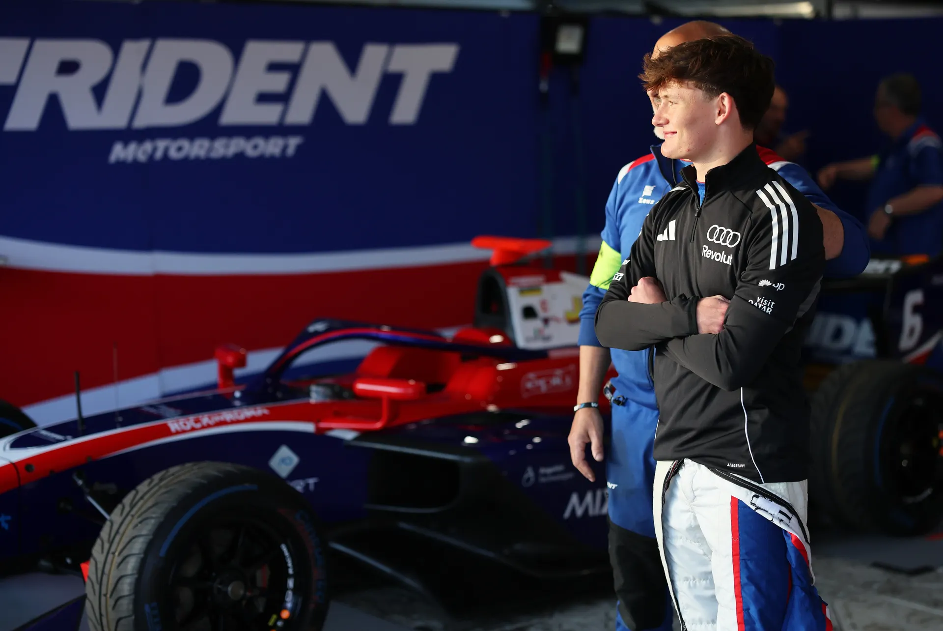 Freddie Slater standing in the garage with arms crossed looking toward the track beside his Formula 3 car