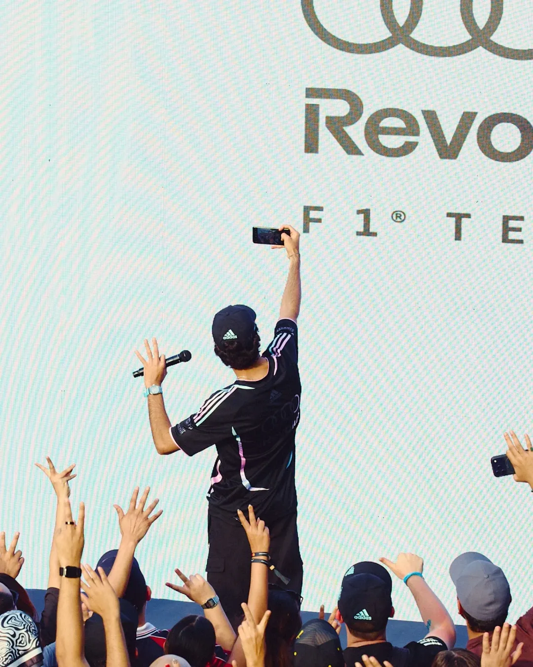 Gabriel Bortoleto stands on stage taking a selfie with the crowd during the Audi Revolut F1® Team fan event at MAPS Backlot in Miami, wearing the special adidas x Audi Revolut F1® Team Miami collection.