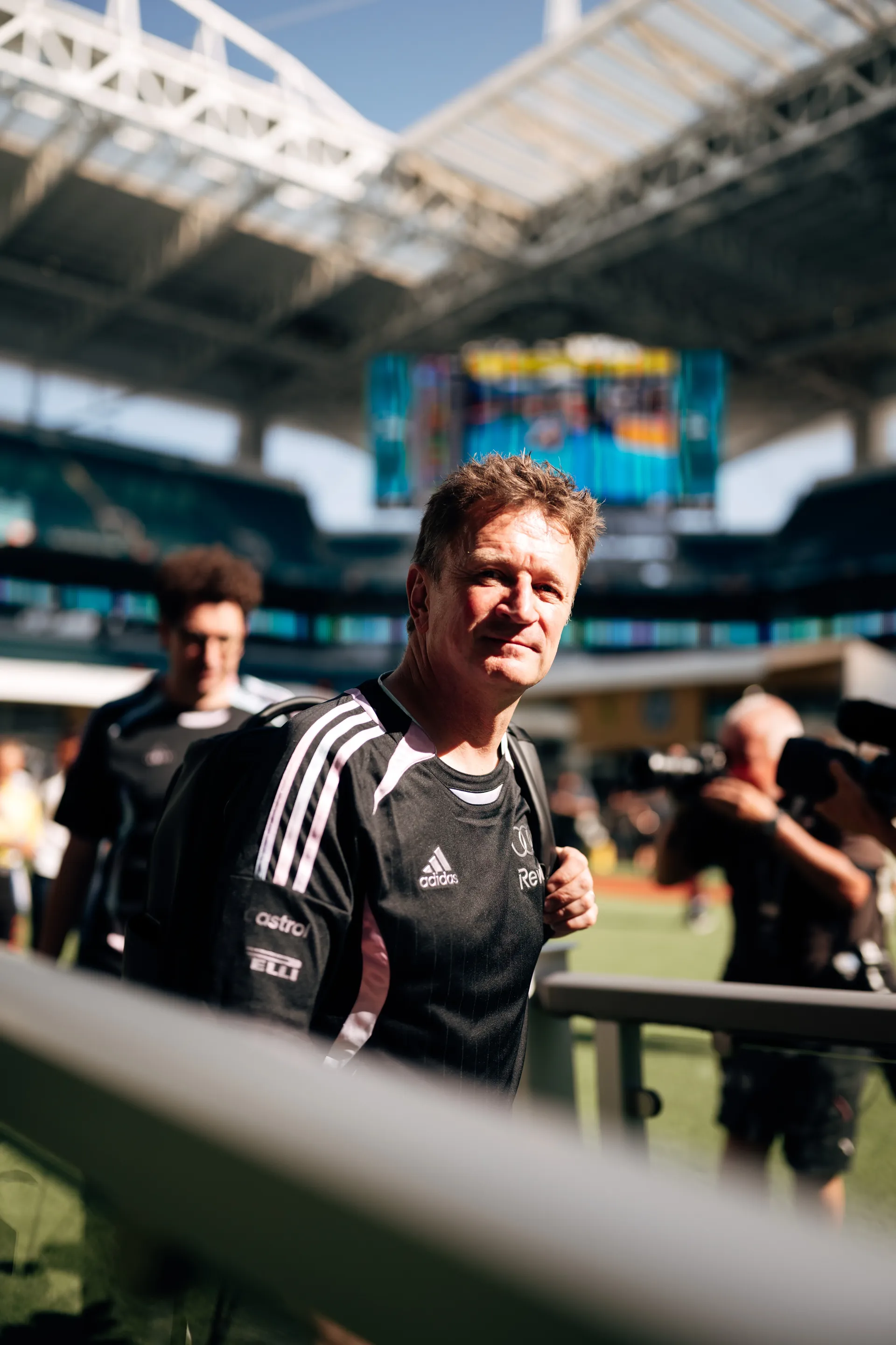 Alan McNish arriving at track in an adidas x Audi Revolut F1® Team shirt, walking across the field inside a stadium while carrying a backpack.