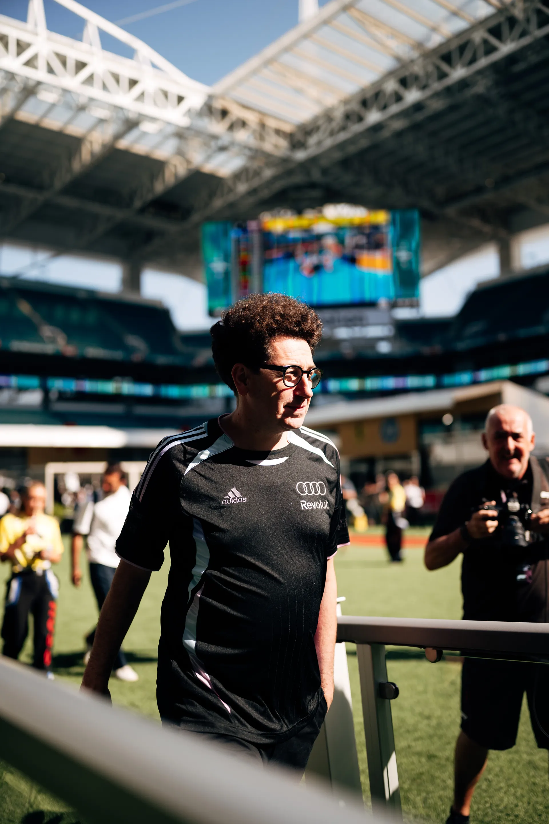 Mattia Binotto arriving at track in an adidas x Audi Revolut F1® Team shirt, walking across the field with the stadium screen in the background.