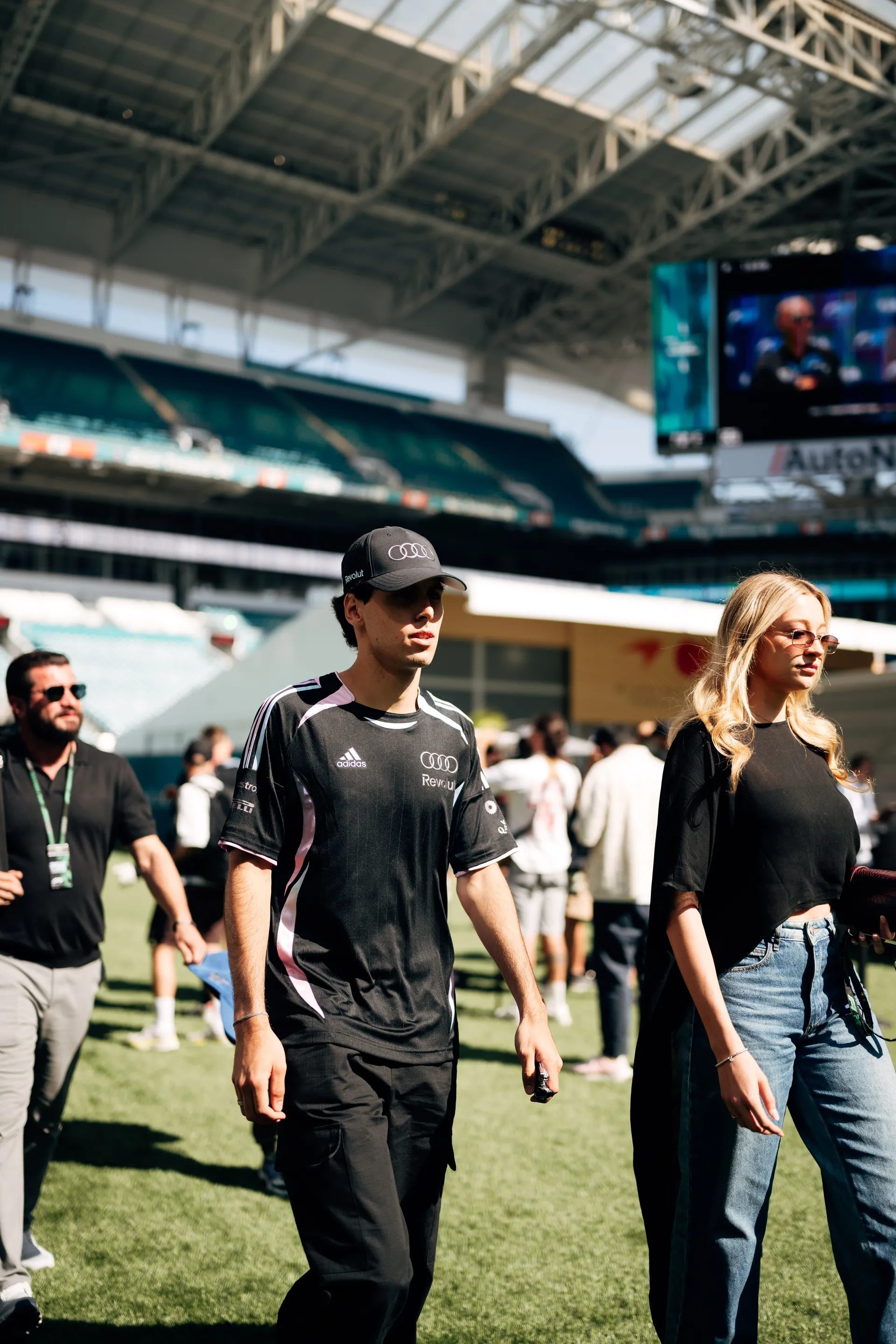 Gabriel Bortoleto arriving at track in an adidas x Audi Revolut F1® Team shirt and cap, walking across the field beside a woman in black and denim.