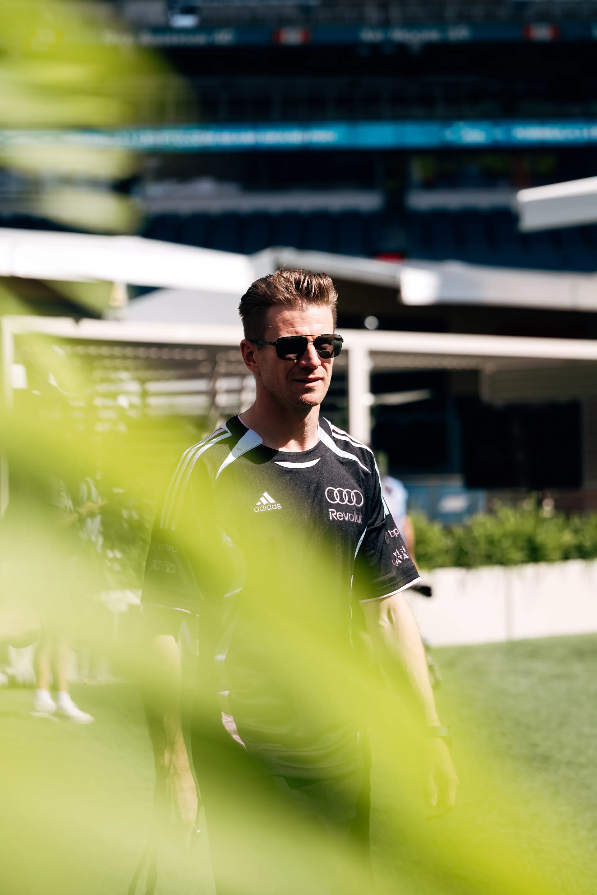 Nico Hulkenberg arriving at track in sunglasses and an adidas x Audi Revolut F1® Team shirt, photographed through blurred green foreground leaves.
