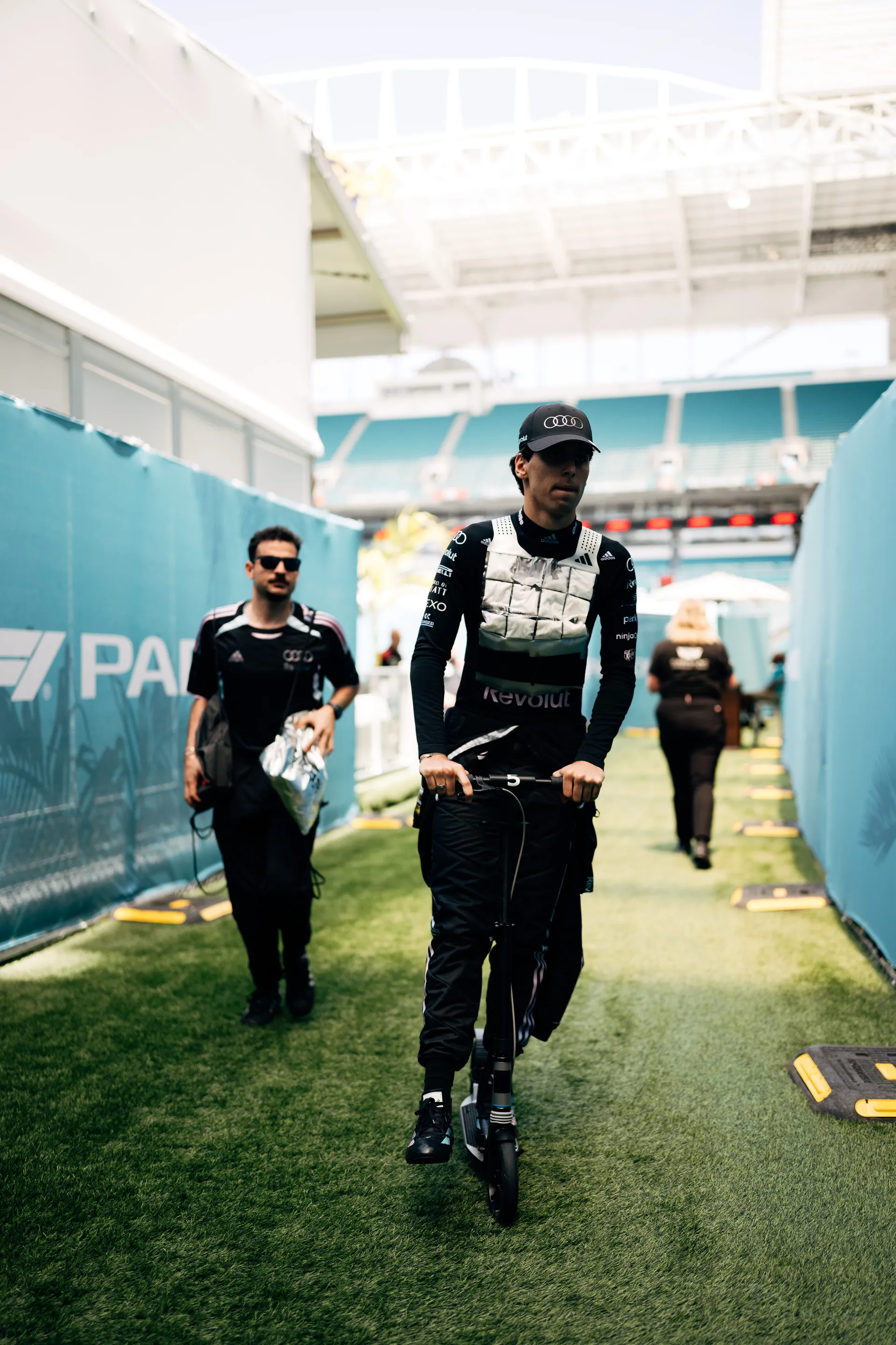 Audi Revolut F1® Team driver Gabriel Bortoleto wearing a black cap and cooling vest rides a scooter through a stadium walkway before track activity.