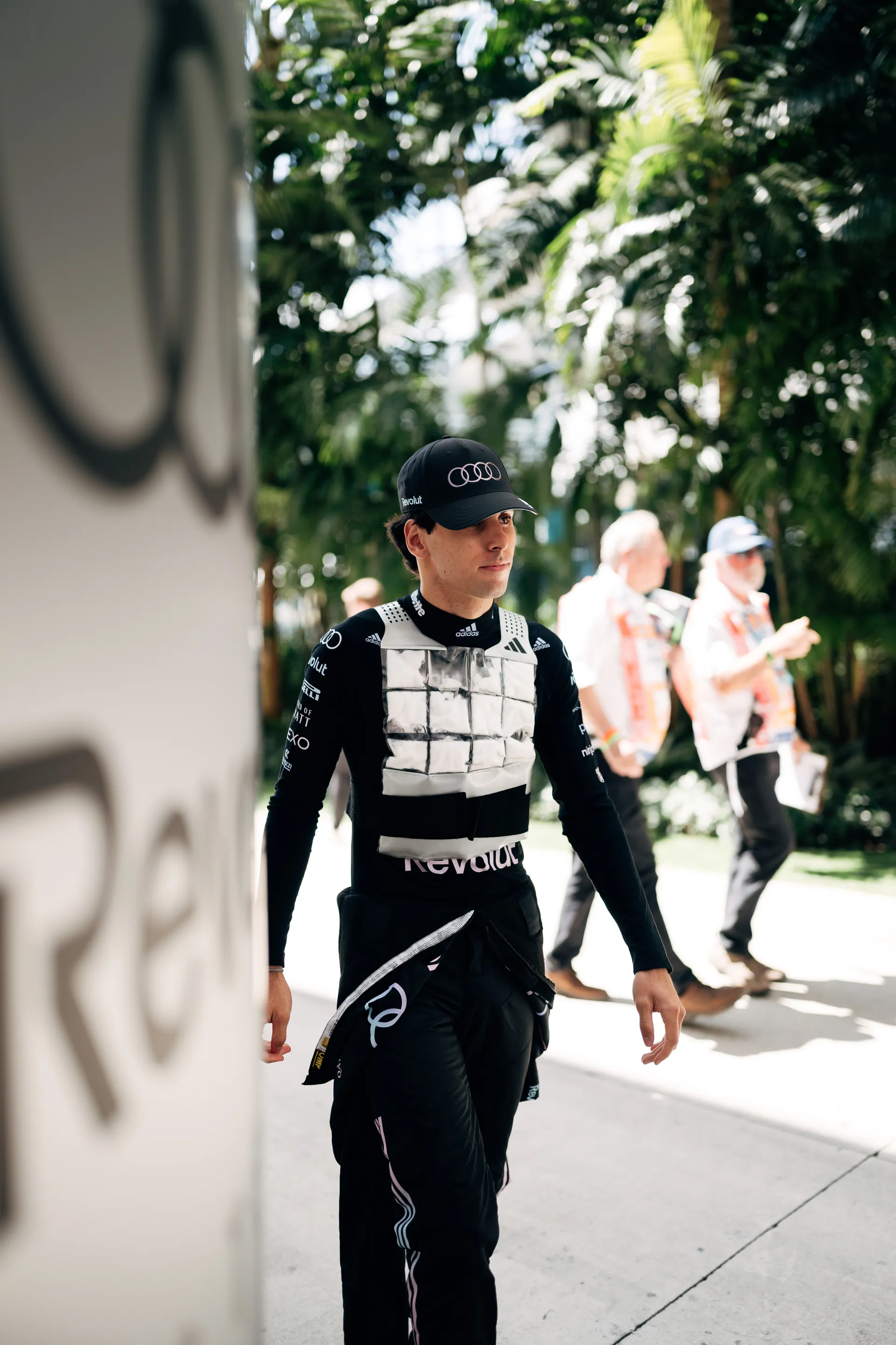 Audi Revolut F1® Team driver Gabriel Bortoleto in racewear and cooling vest walks past palm trees in the paddock at the Miami venue.