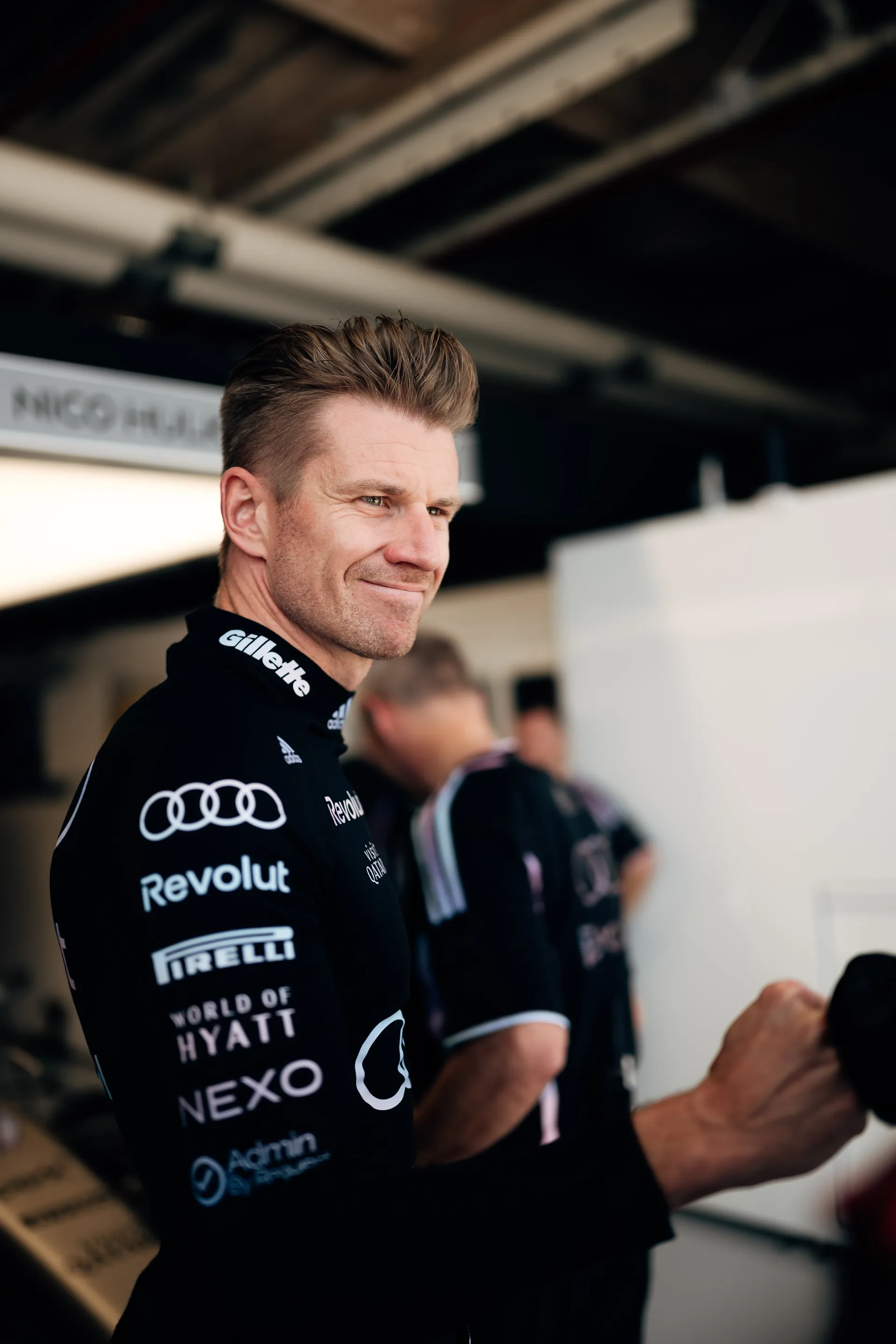 Audi Revolut F1® Team driver Nico Hulkenberg in black race base layer smiles in the garage area with the Nico Hulkenberg bay sign blurred in the background.