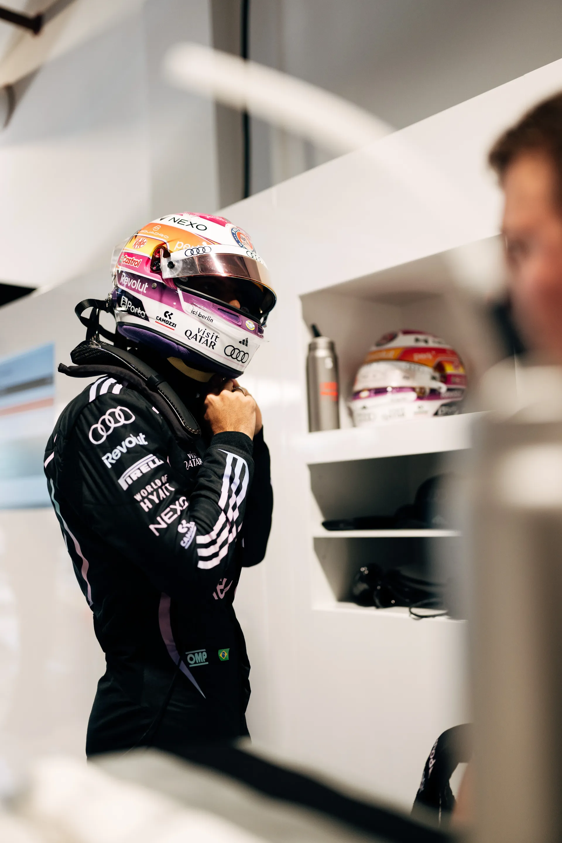 Audi Revolut F1® Team driver Gabriel Bortoleto wearing the special Miami  helmet adjusts his race gear beside white storage shelves in the garage.