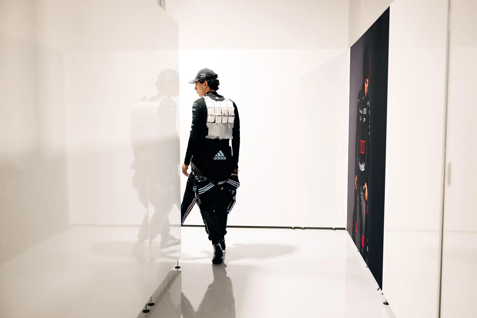 Gabriel Bortoleto walks through a bright white paddock corridor wearing his Audi Revolut F1® Team race suit, cap, and a silver cooling vest, carrying the upper part of his suit at his waist.
