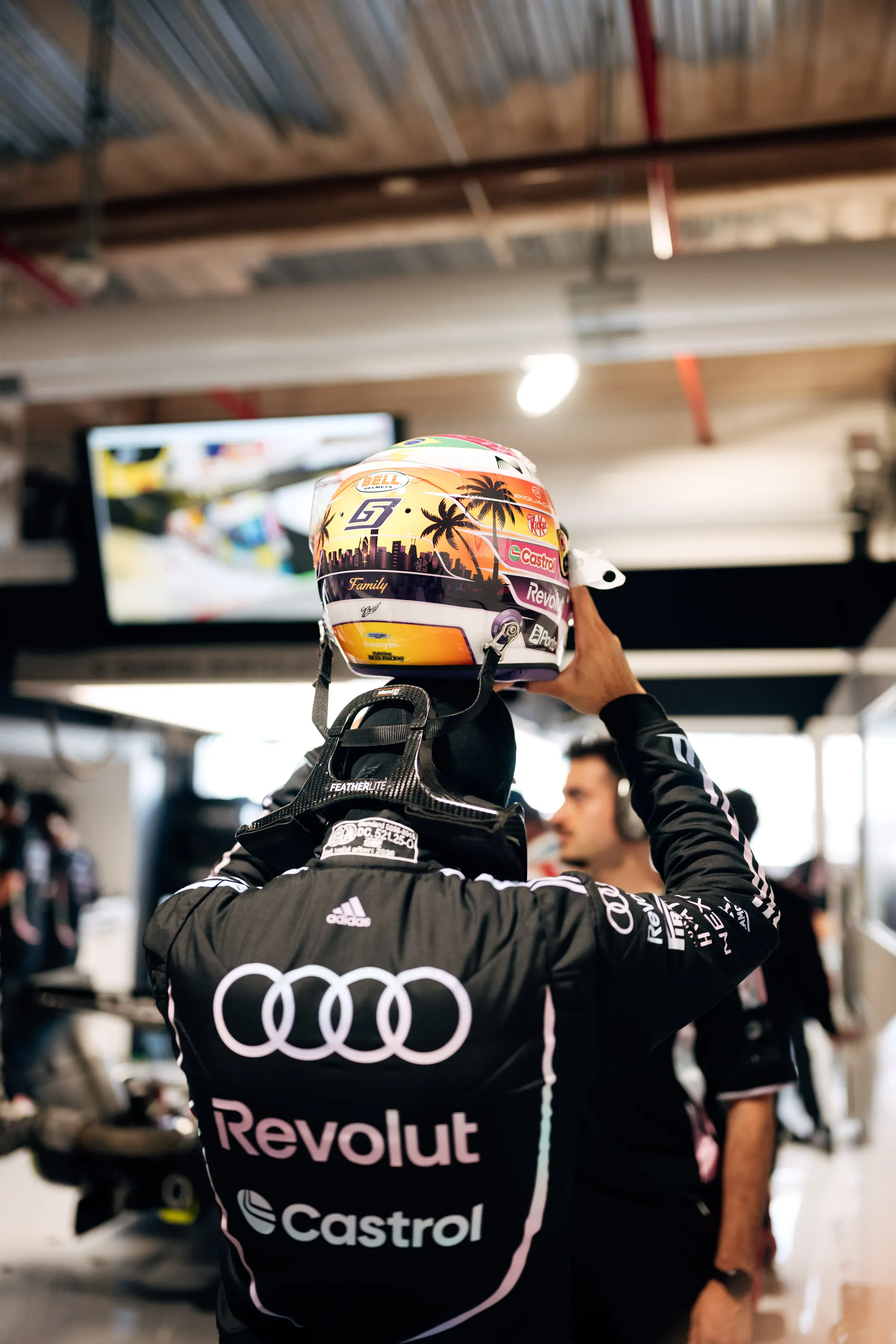 Rear view of Gabriel Bortoleto in the Audi Revolut F1® Team race suit as he lifts his special Miami edition helmet in the garage, with the skyline and palm tree artwork visible on the back.