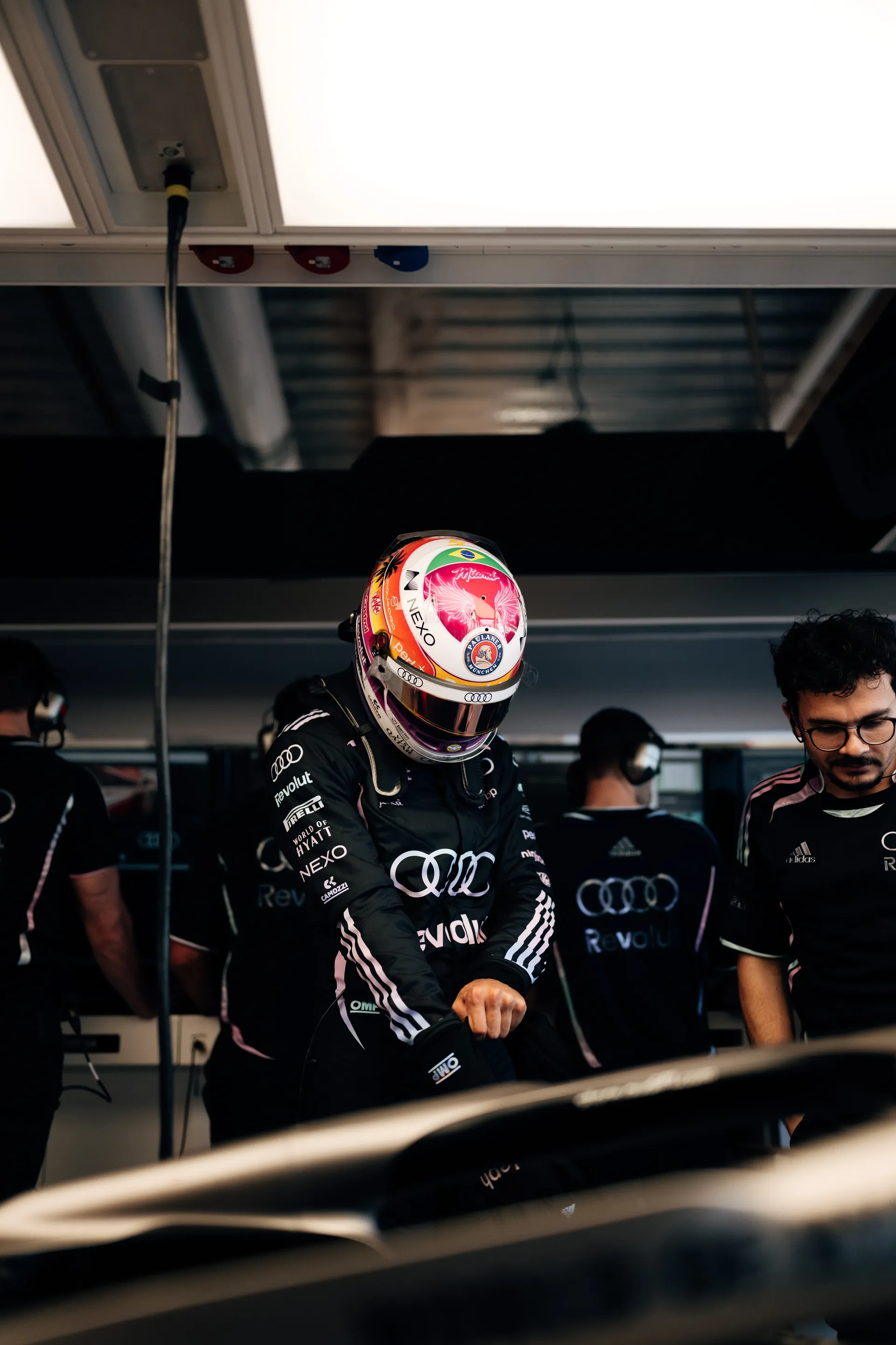 Gabriel Bortoleto stands in the Audi Revolut F1® Team garage wearing his special Miami edition helmet and race suit while pulling on a glove beside team members.