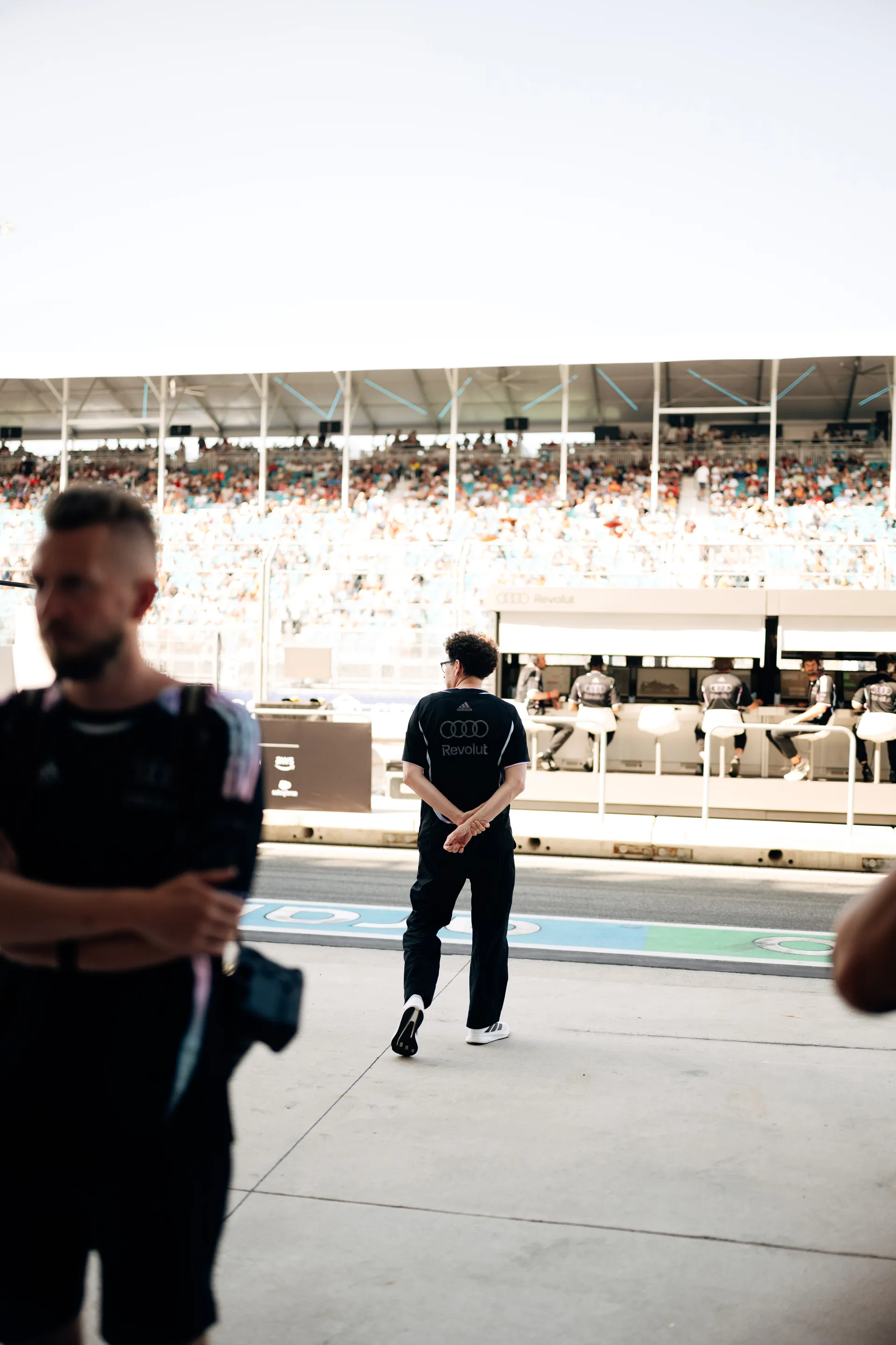Mattia Binotto walks along the Miami pit lane in Audi Revolut F1® Team kit, framed by blurred figures in the foreground and grandstands full of spectators in the background.