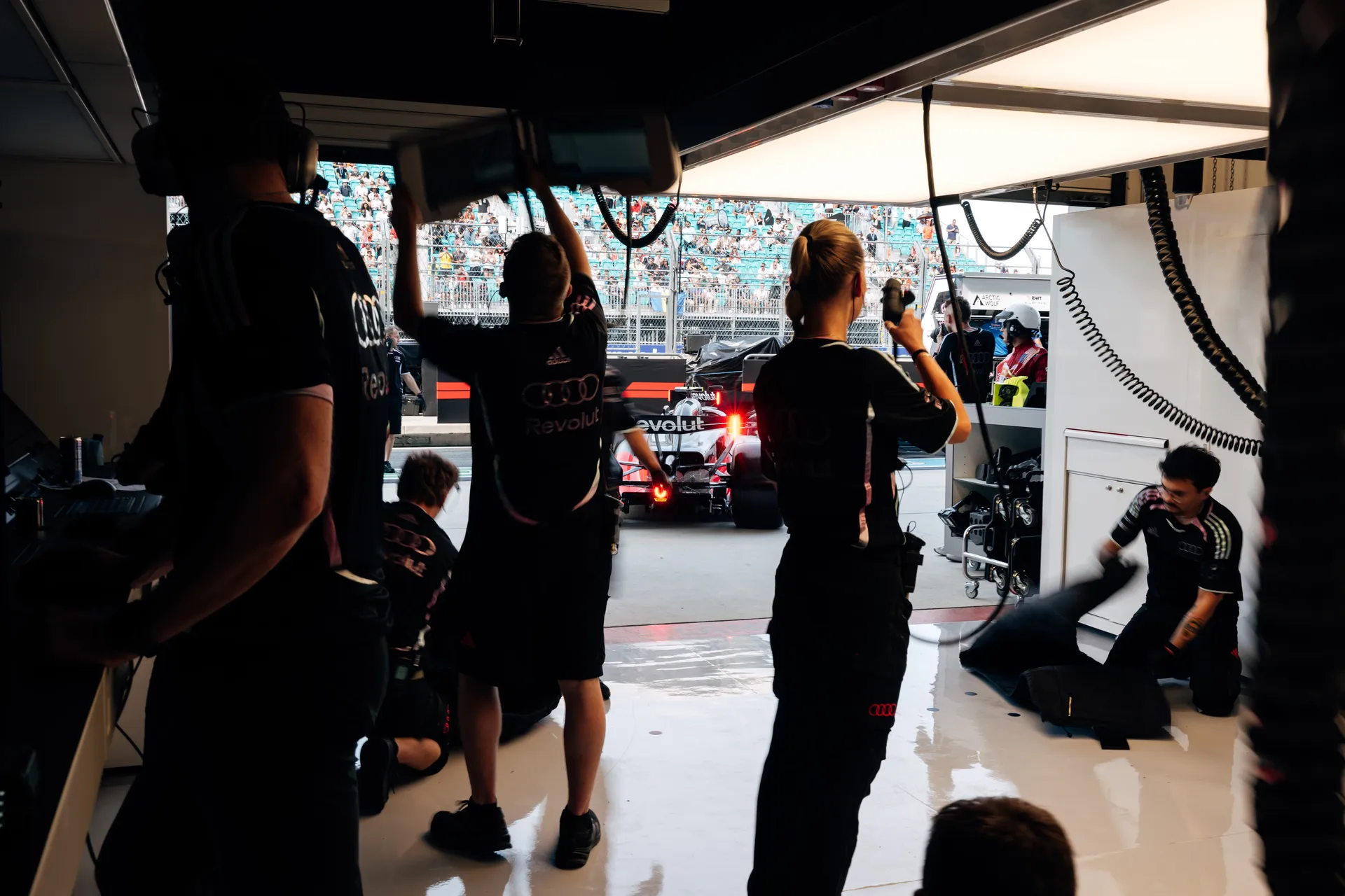 The Audi Revolut F1® Team pit crew prepares in the garage as the Formula 1 car exits toward the pit lane, with overhead equipment raised and grandstands visible outside.