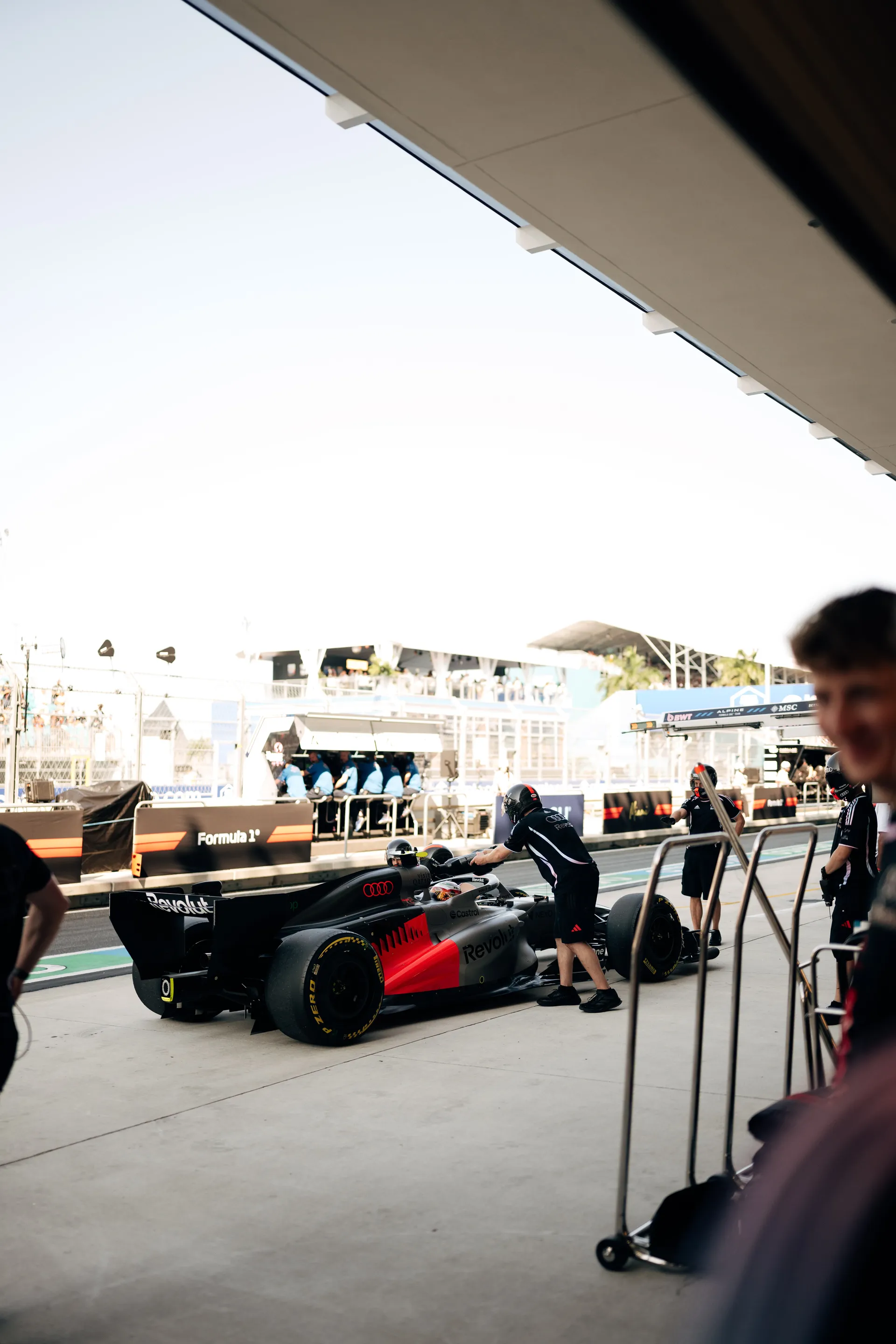 The Audi Revolut F1® Team pit crew prepares in the garage as the Formula 1 car exits toward the pit lane, with overhead equipment raised and grandstands visible outside.