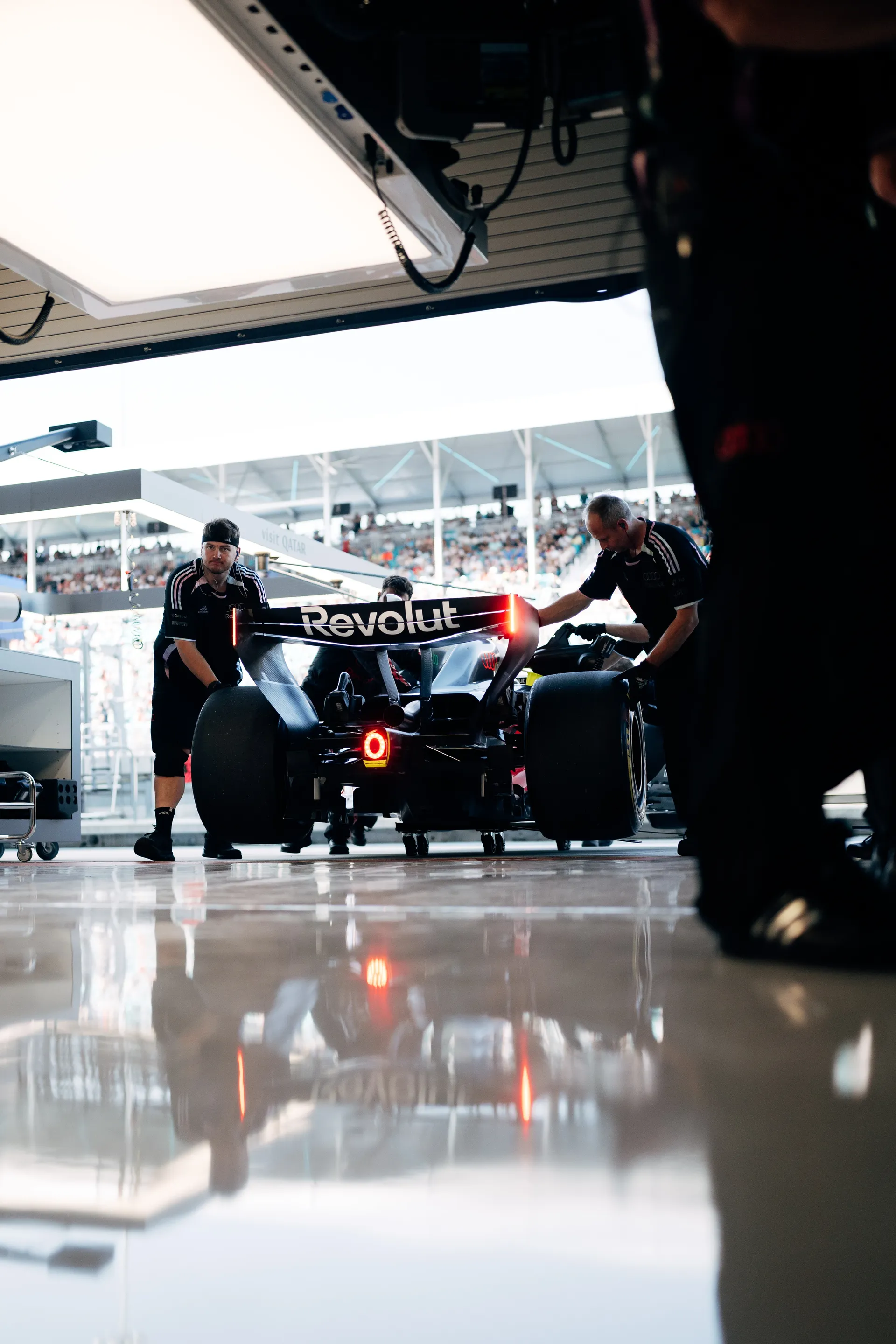 Rear view of the Audi Revolut F1® Team Formula 1 car being guided into the garage by crew members, with the rear light glowing and reflections visible on the polished floor.