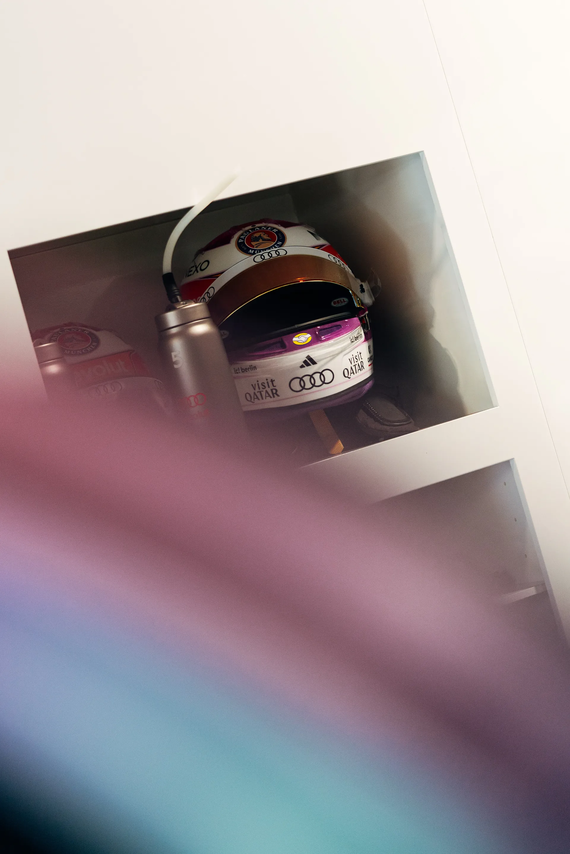 Racing helmet resting in an open garage shelf beside a metal water bottle, photographed through soft pink and blue blur in the foreground.