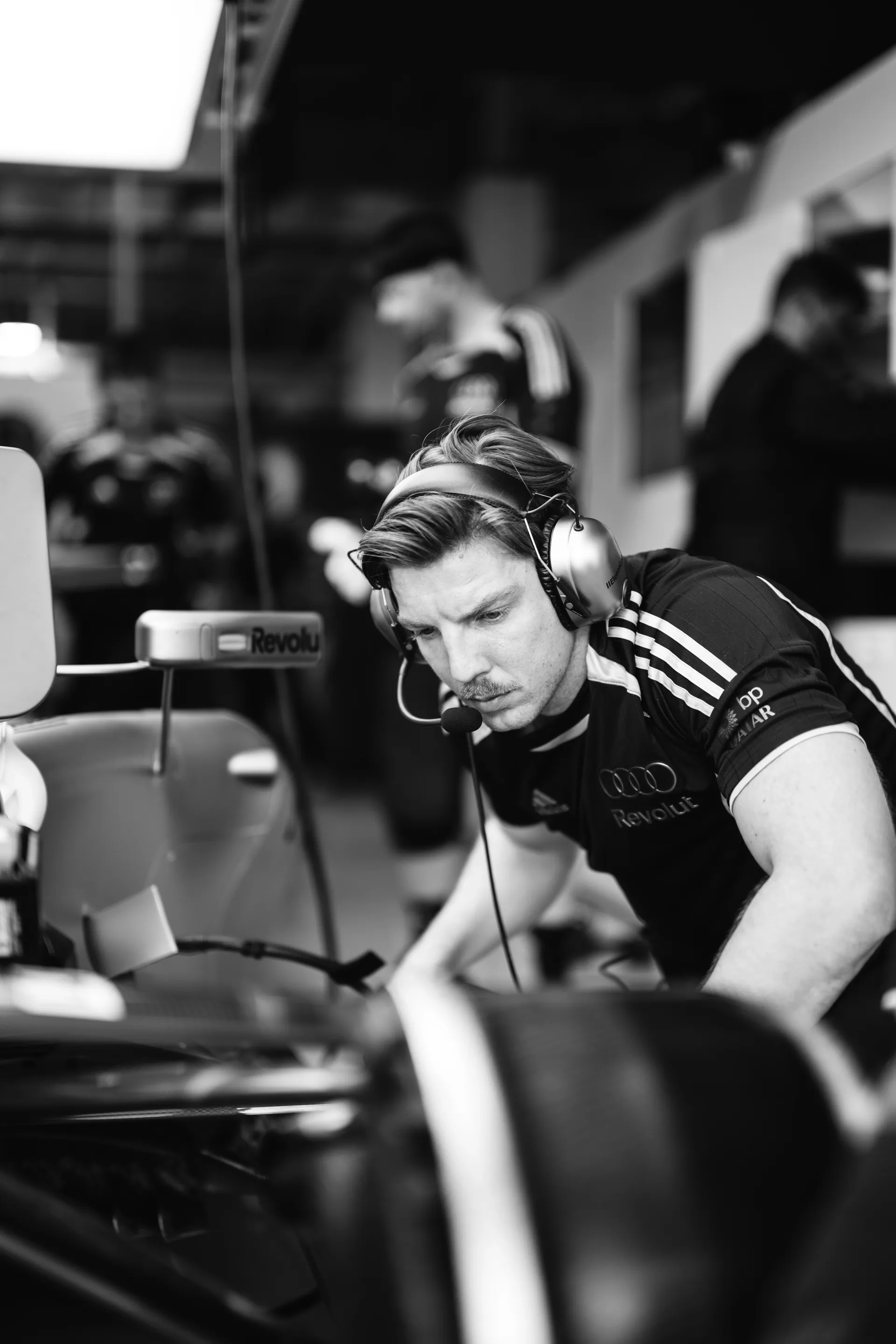 Black-and-white close-up of an engineer wearing a headset, focused on the car setup inside the garage.