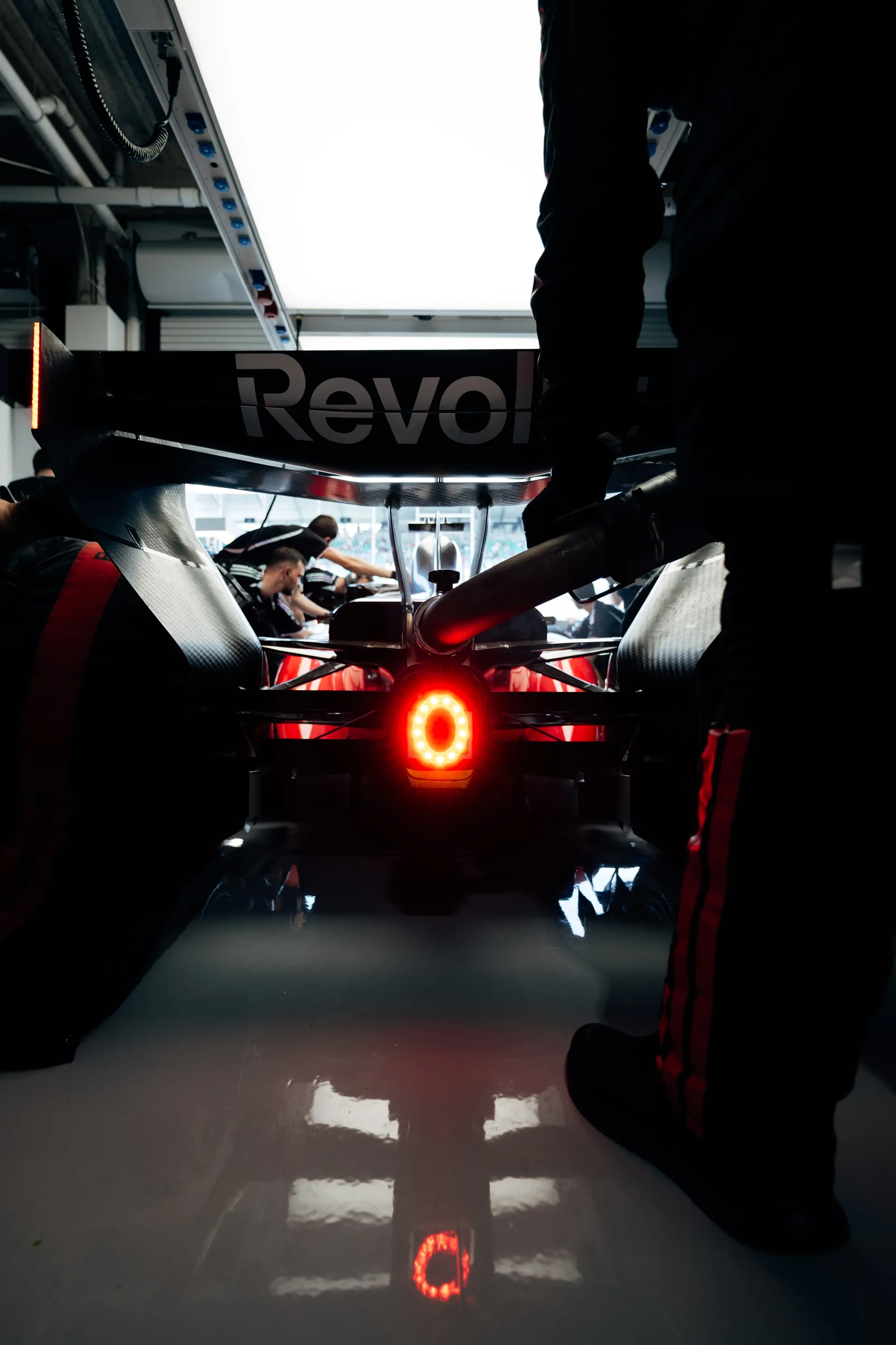 Rear view of the Formula 1 car inside the garage, framed by crew members and the glowing red rear light reflected on the polished floor.