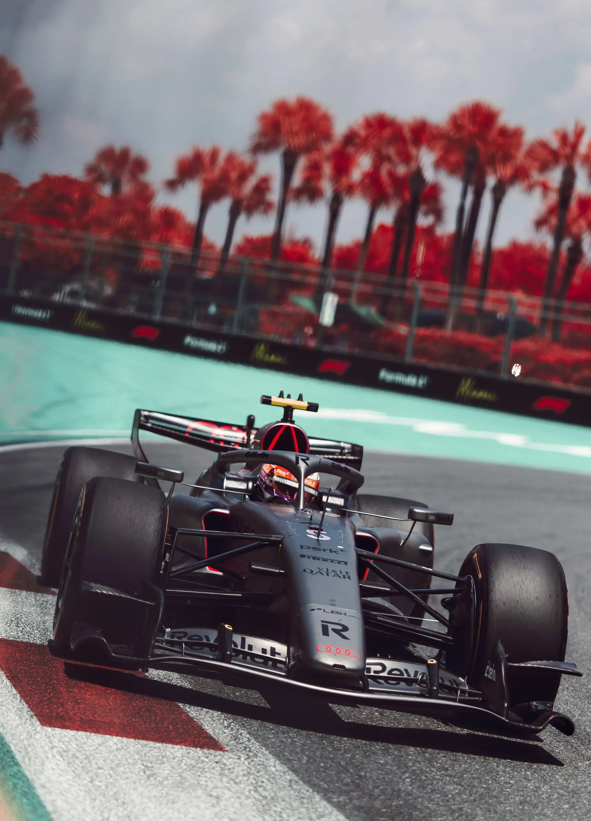 Audi Revolut F1® Team car cornering at Miami circuit with orange helmet visible and stylized red palm trees in background