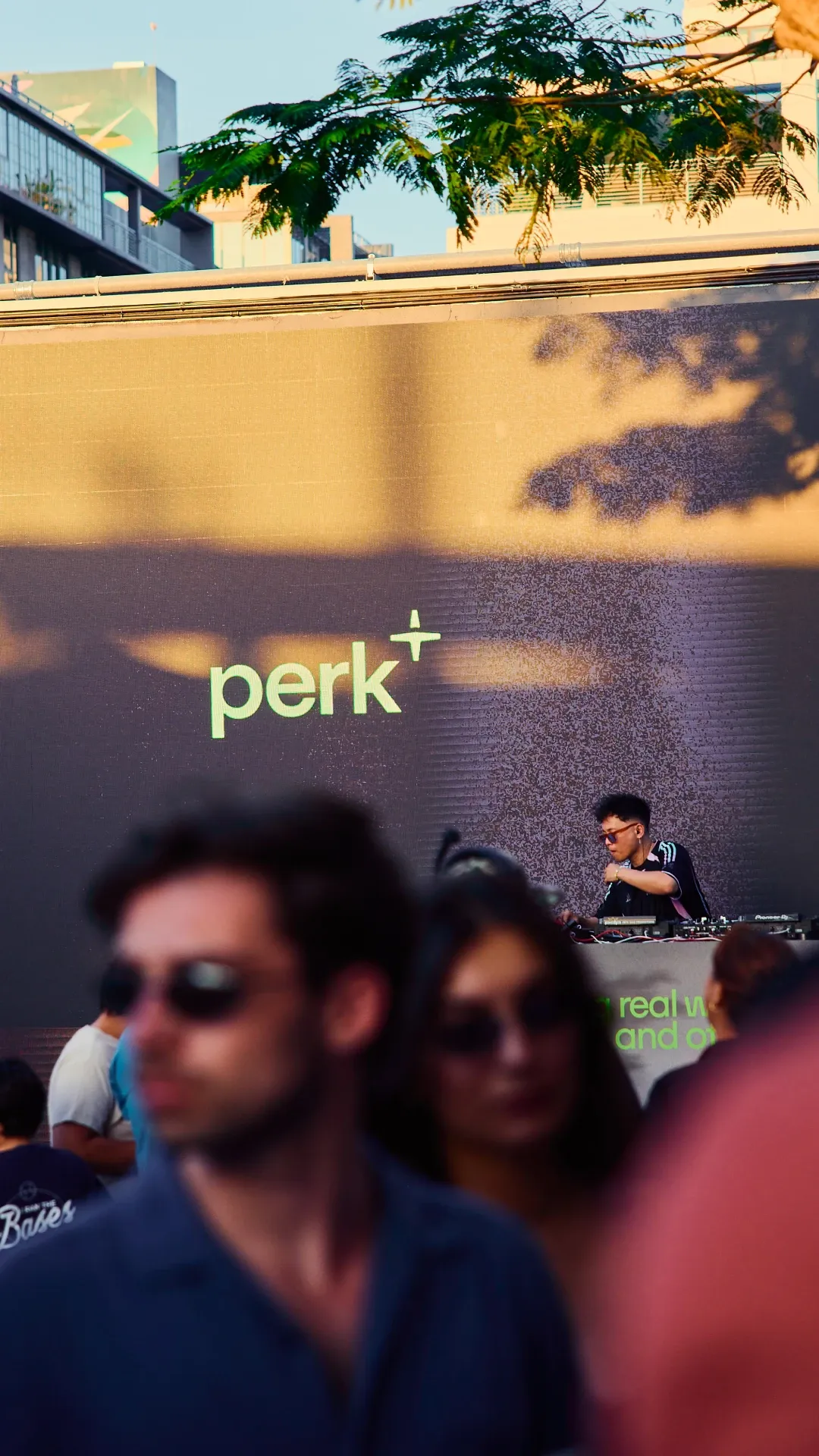 DJ performing on stage beneath a “perk+” branded screen while attendees watch from the crowd