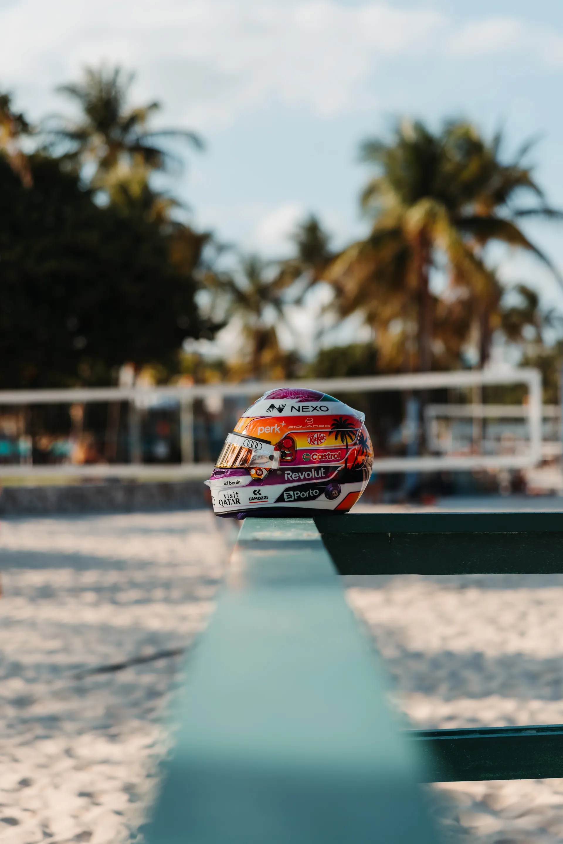 Side profile of Gabriel Bortoleto’s special Miami edition race helmet resting on a turquoise beach bench, with sand, palm trees, and a blurred volleyball net behind it.