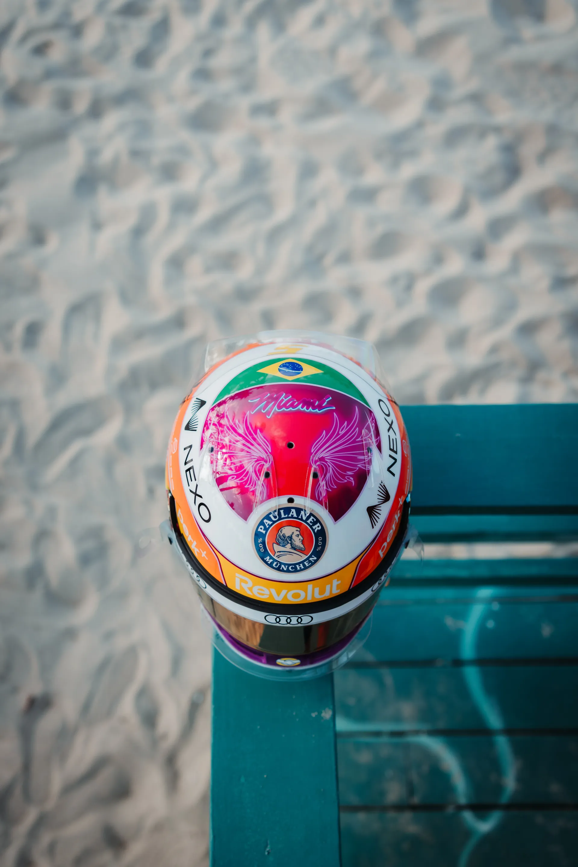 Top down view of Gabriel Bortoleto’s special Miami edition race helmet on a turquoise beach bench, showing the Brazil flag and pink Miami artwork on the crown.