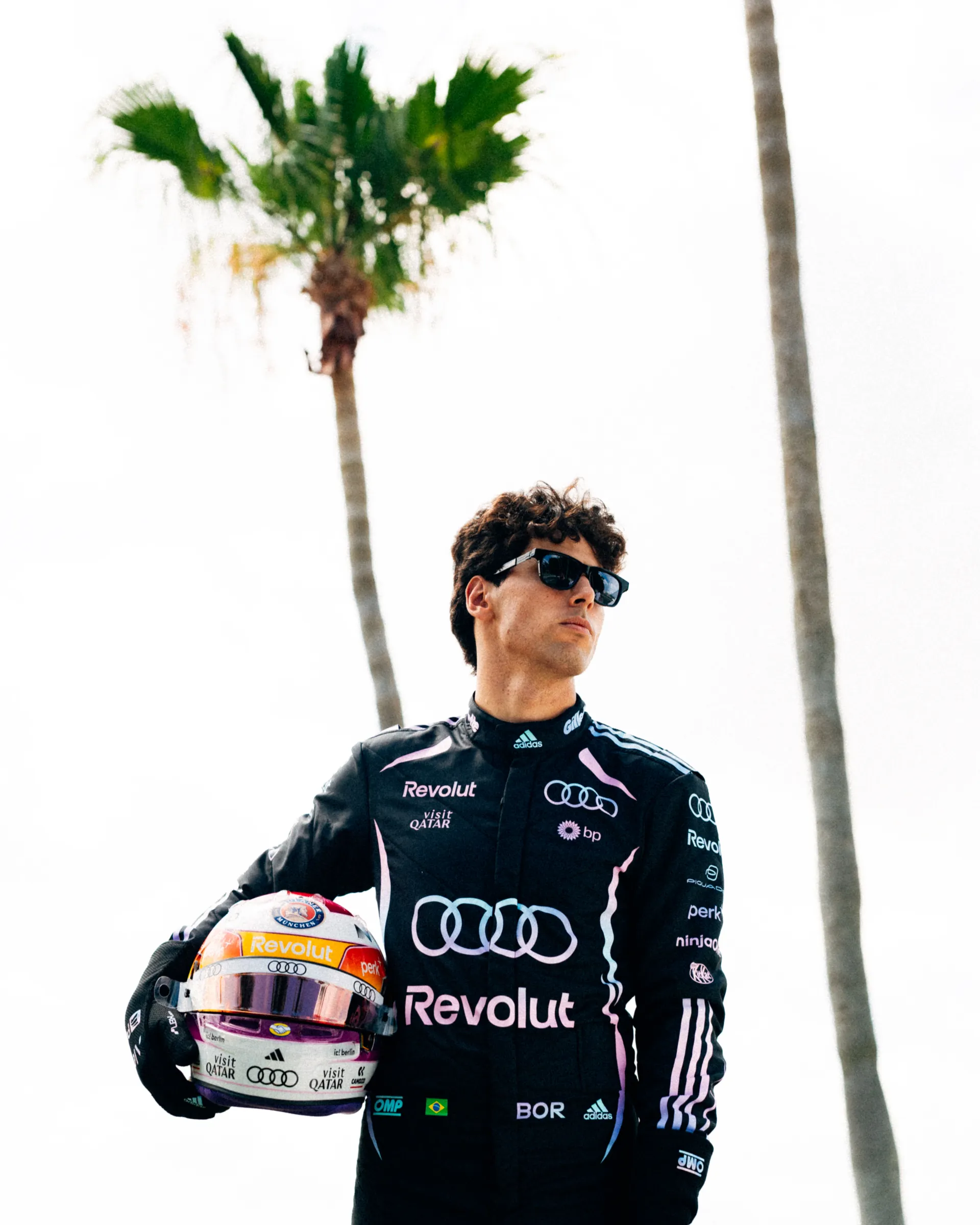Gabriel Bortoleto in the Audi Revolut F1® Team Miami race suit holding his special Miami edition helmet beneath tall palm trees.