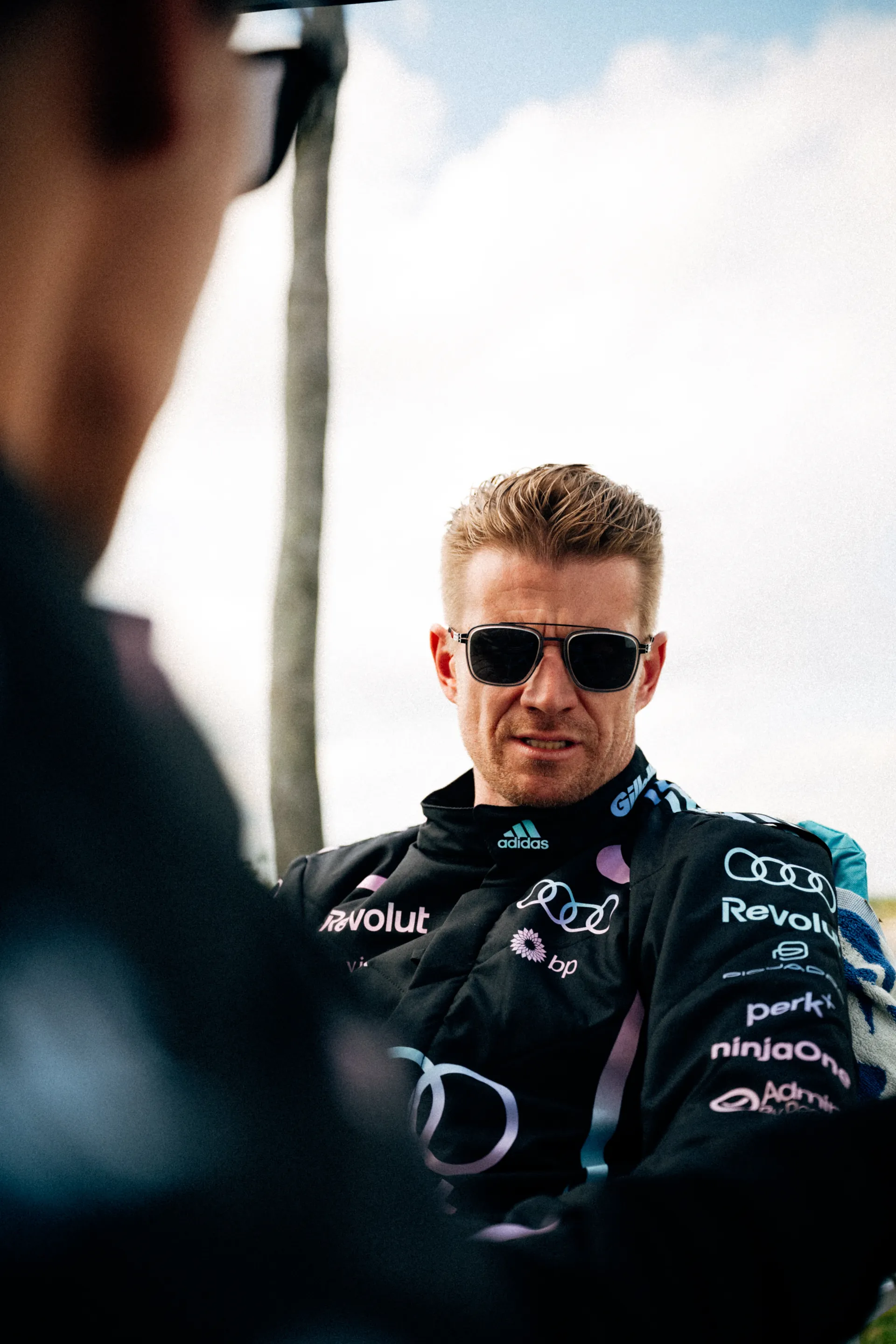 Nico Hulkenberg sits outdoors in the Audi Revolut F1® Team Miami race suit and dark ic! berlin sunglasses, looking ahead with palm trees and bright sky behind him.