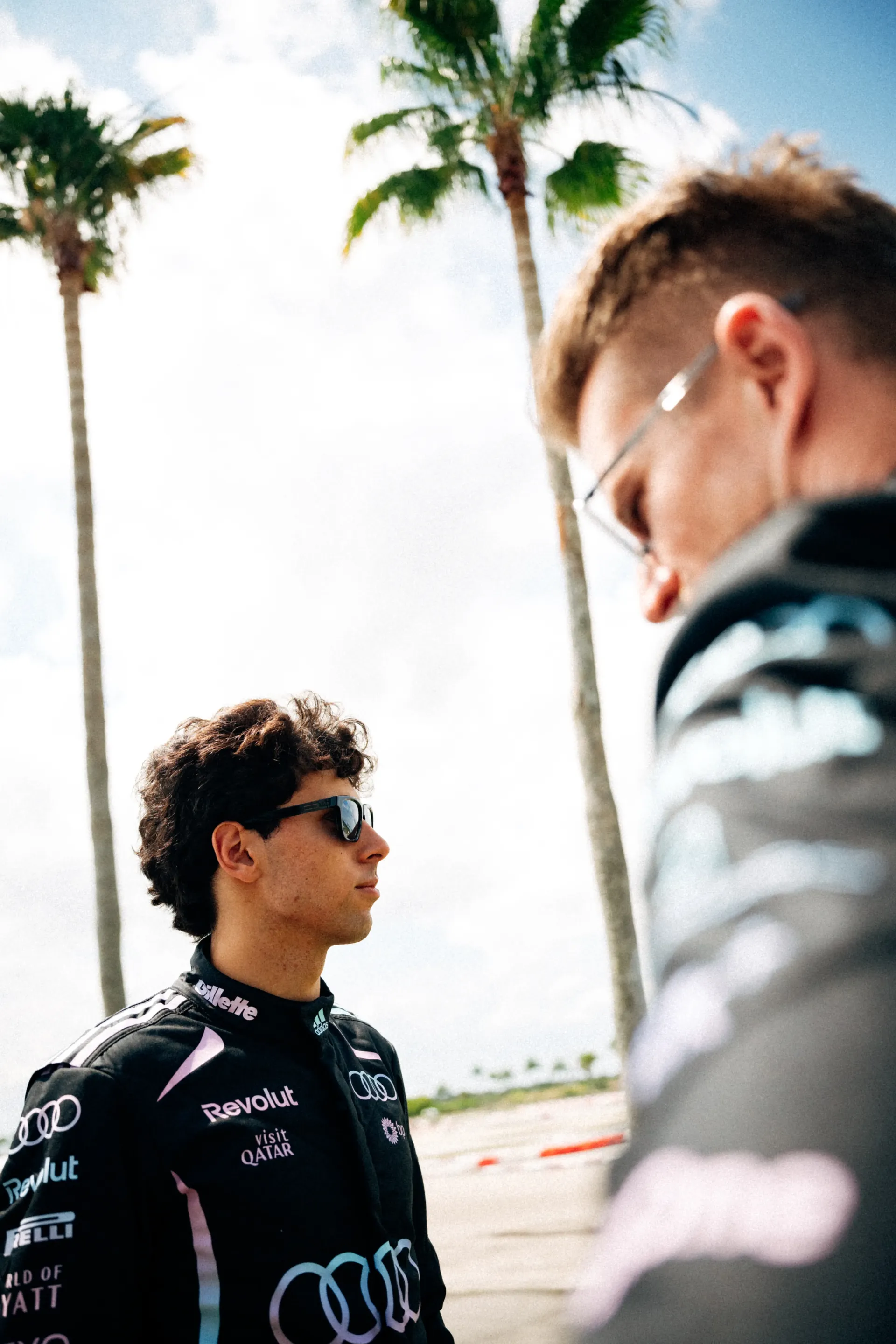 Gabriel Bortoleto stands in profile wearing the Audi Revolut F1® Team Miami race suit and black ic! berlin sunglasses, while Nico Hulkenberg appears blurred in the foreground with palm trees in the background.