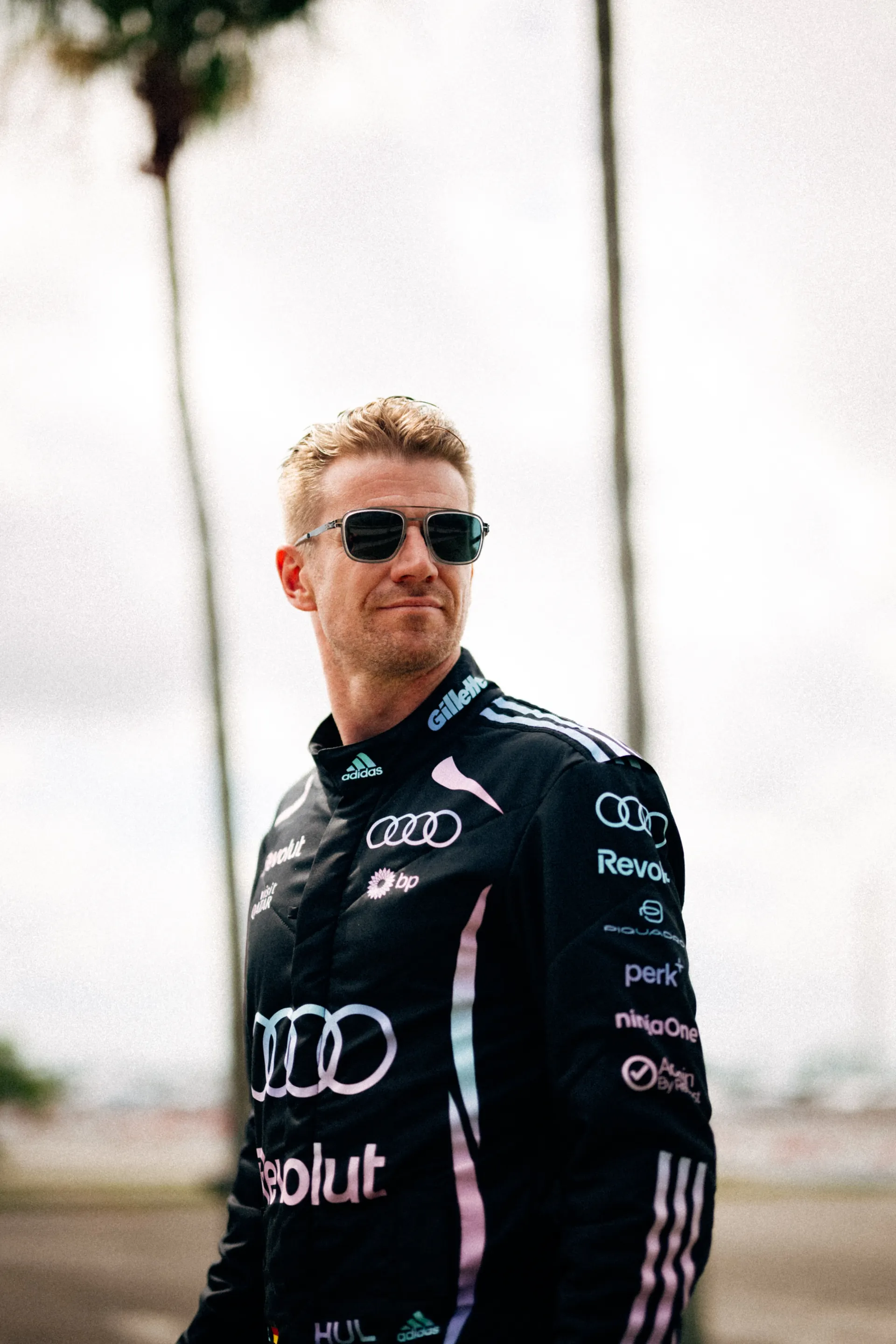Nico Hulkenberg stands outdoors in the Audi Revolut F1® Team Miami race suit and dark ic! berlin sunglasses, with tall palm trees rising behind him.