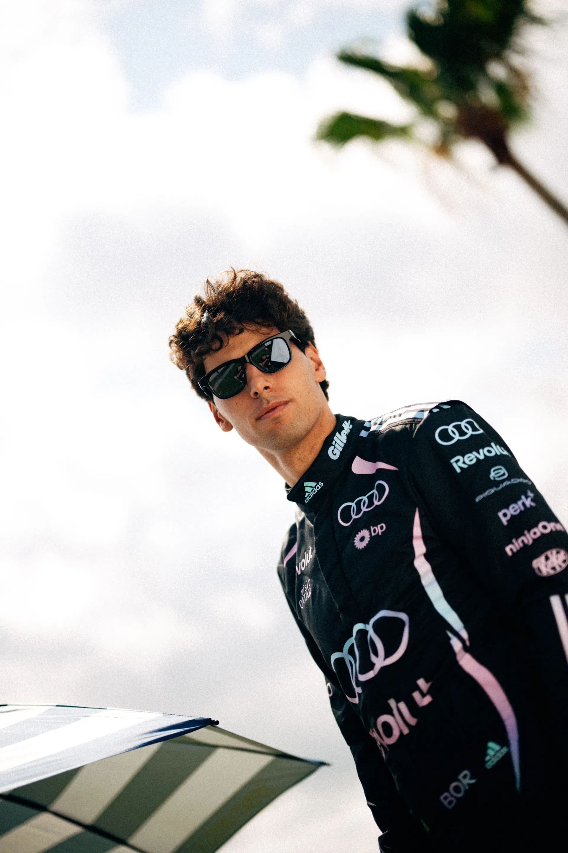 Gabriel Bortoleto is photographed from a low angle in the Audi Revolut F1® Team Miami race suit and black ic! berlin sunglasses, with a striped parasol and palm tree above him.