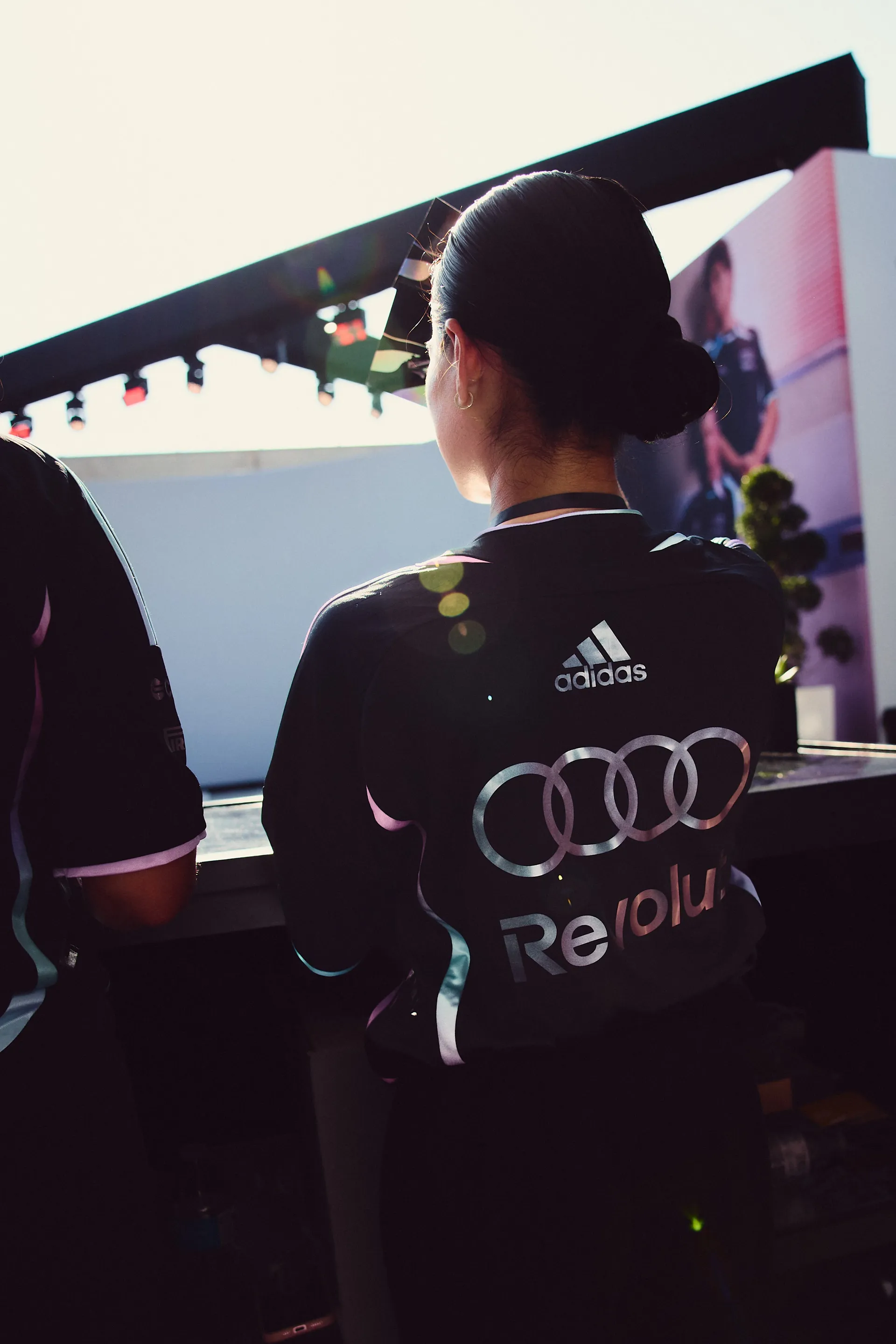 Event attendee wearing Audi Revolut F1® Team x adidas jersey seen from behind, watching stage content in a sunlit Miami setting.