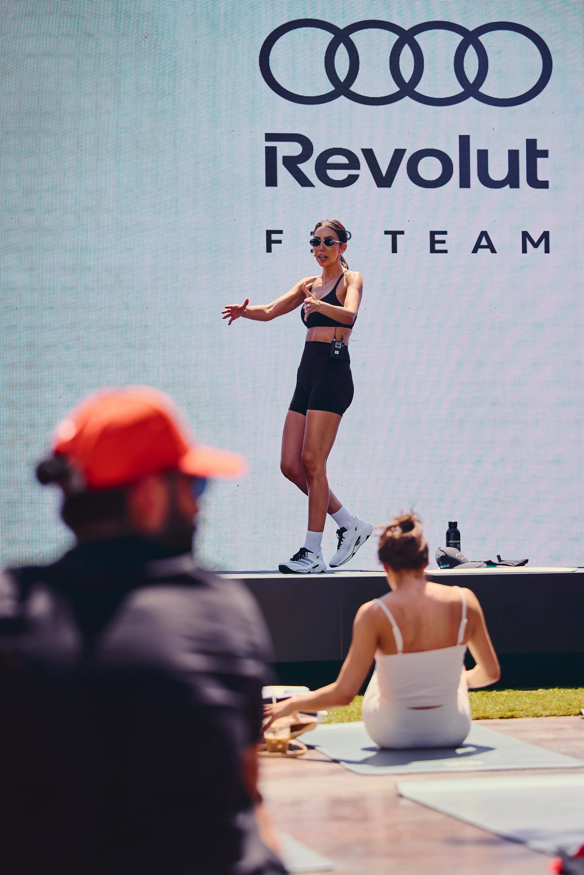 Female trainer leading a workout session on stage at the Audi Revolut F1® Team activation, engaging participants in front of branded backdrop.