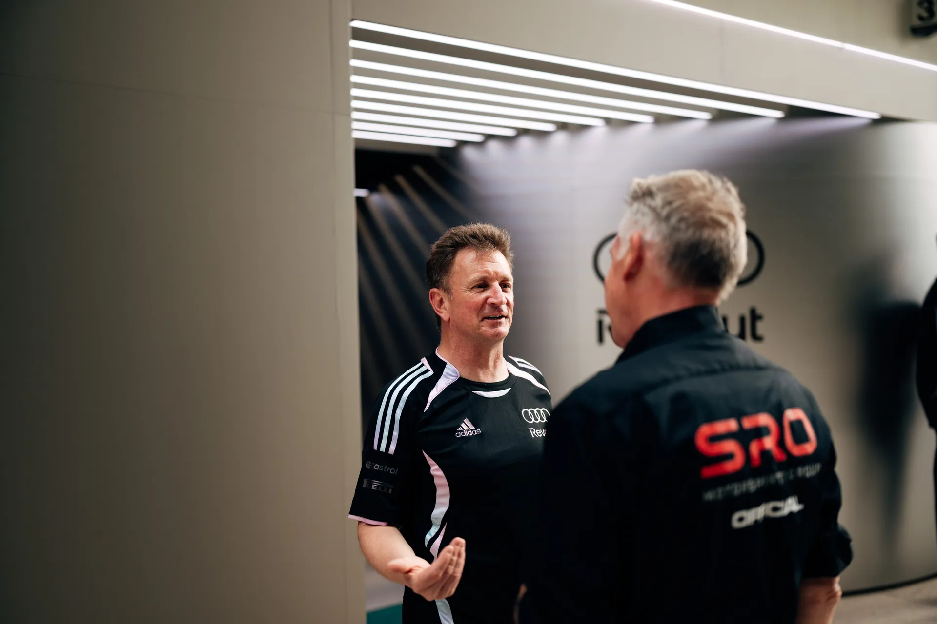 Alan McNish of Audi Revolut F1® Team in conversation with an SRO official inside a lit paddock corridor