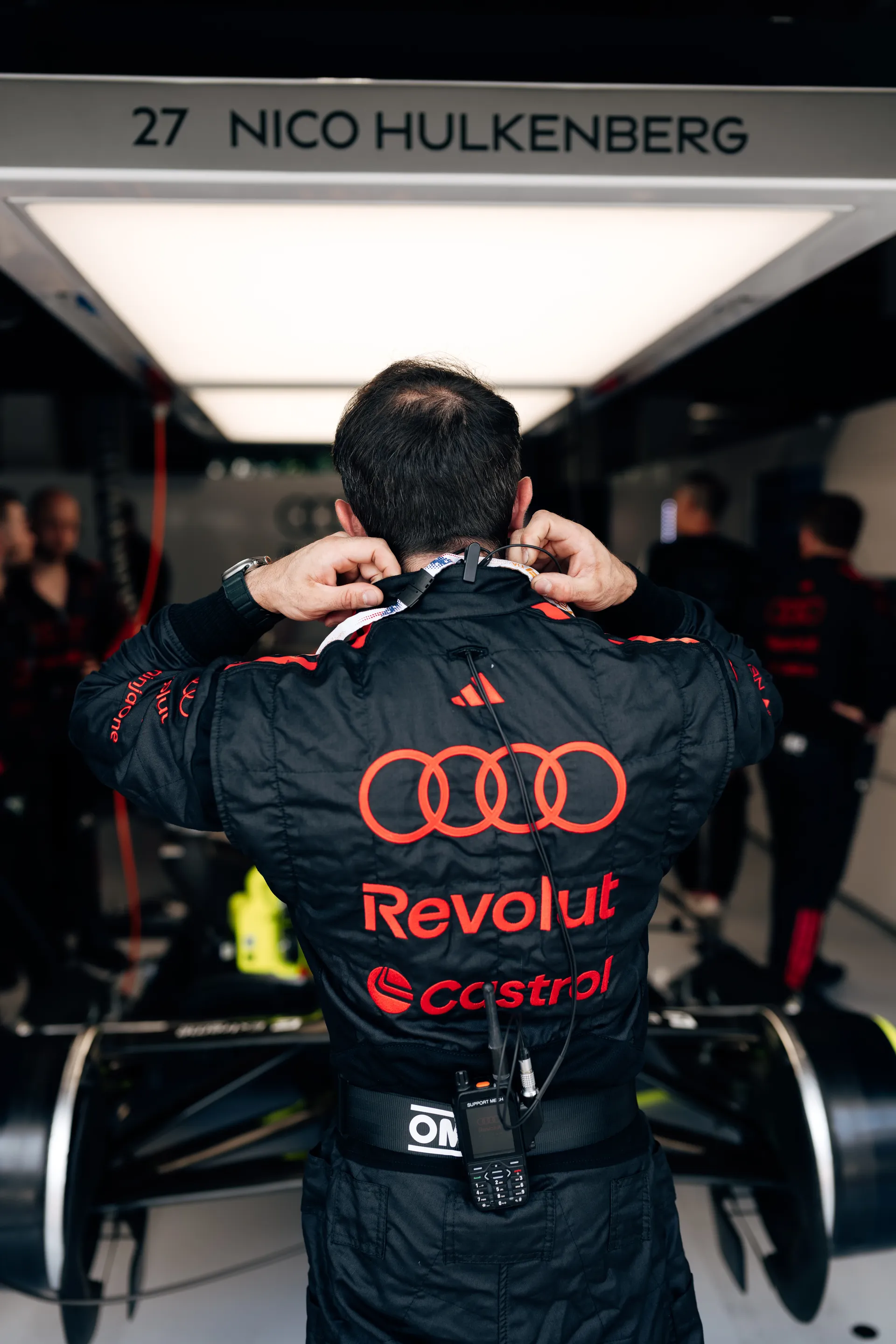 Rear view of a driver beneath a “Nico Hulkenberg” garage sign adjusting his race suit in the Audi Revolut F1® Team pit area