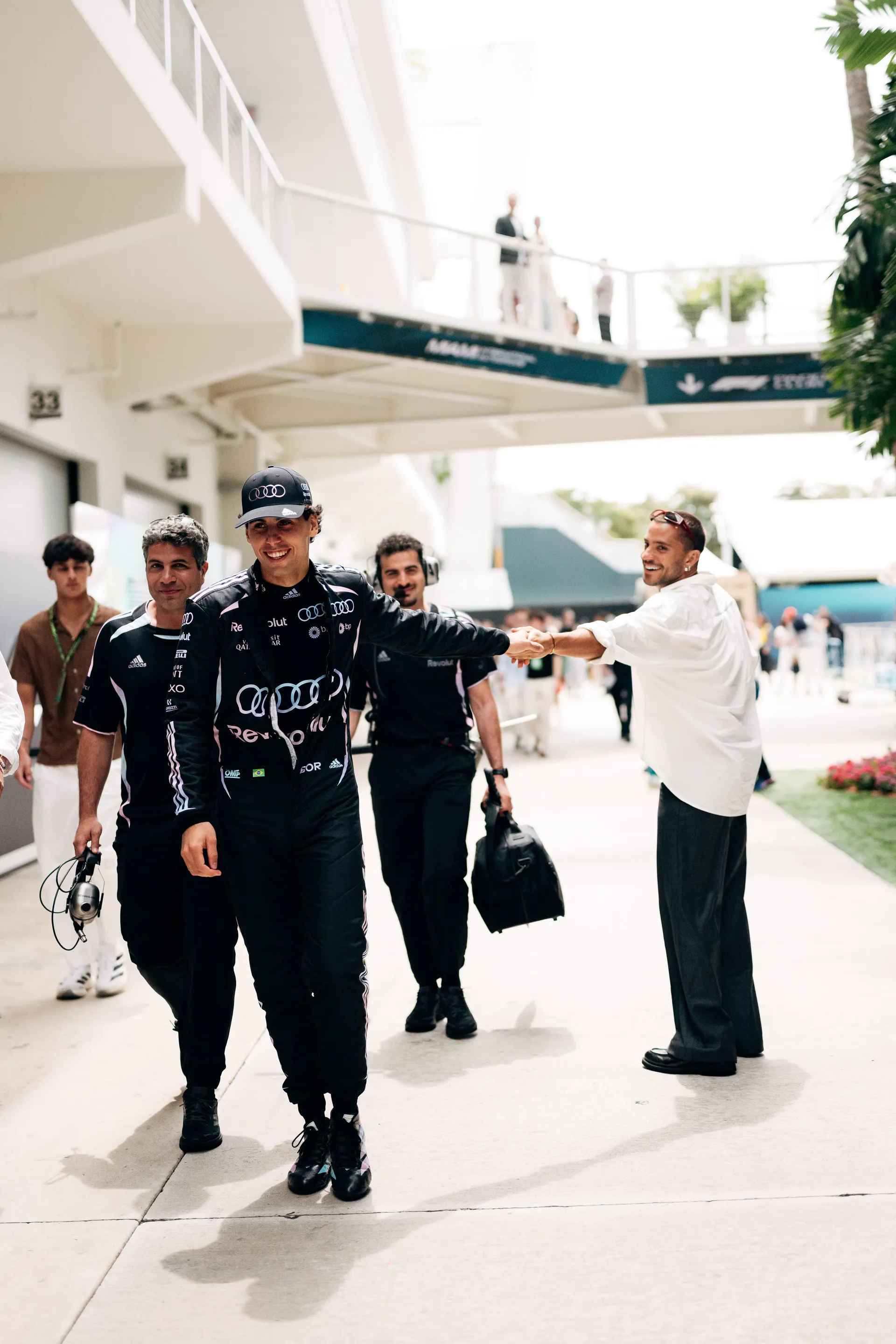 Gabriel Bortoleto walking through the paddock with Audi Revolut F1® Team members, greeting a person with a handshake while heading toward the garage area.
