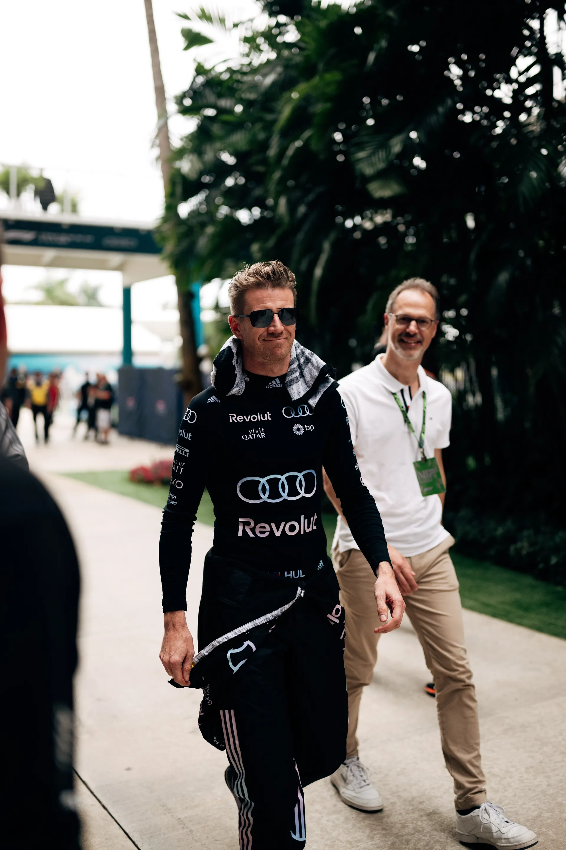 Nico Hulkenberg walking through the paddock in Audi Revolut F1® Team race suit, wearing sunglasses and carrying gear ahead of a session.