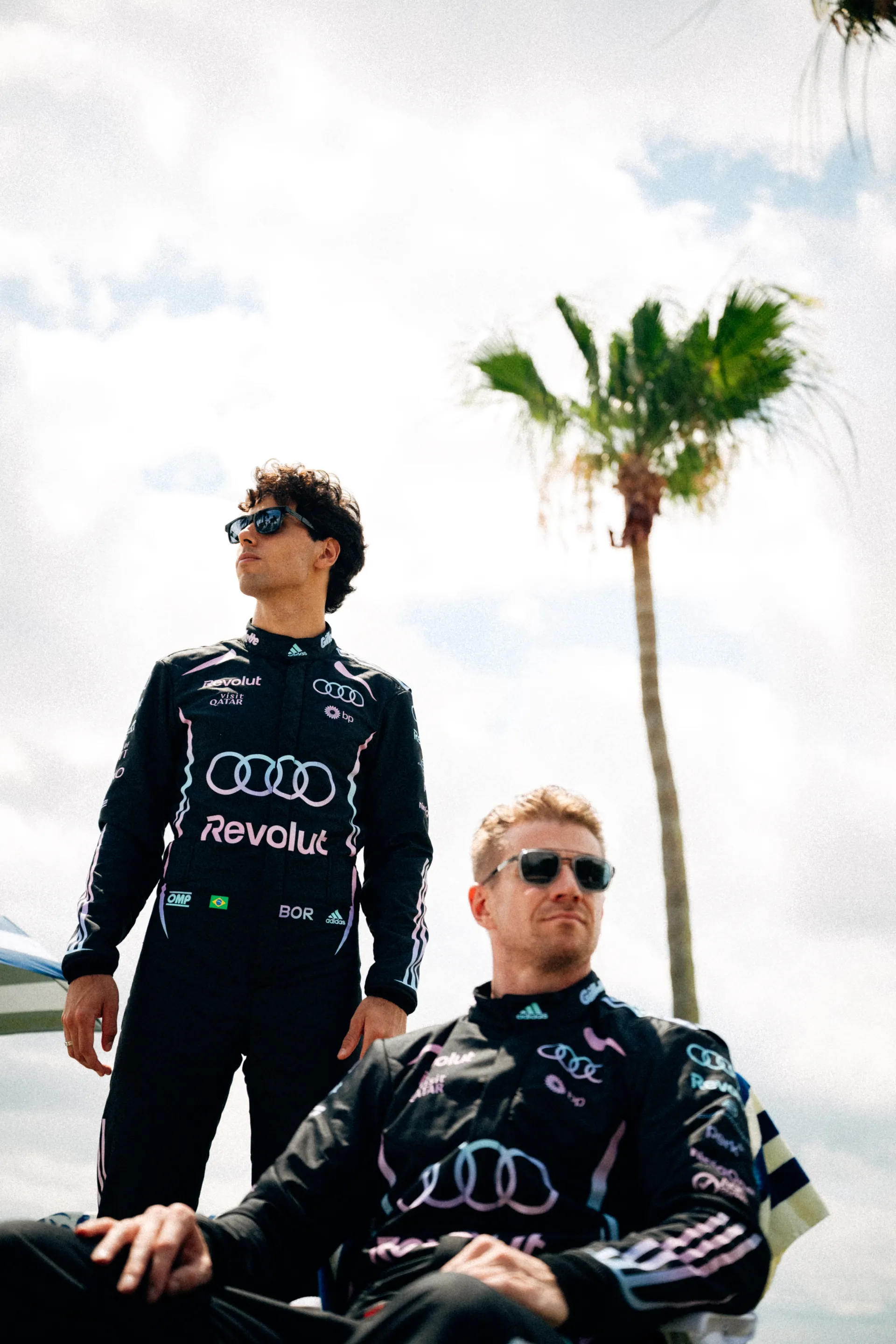 Gabriel Bortoleto stands while Nico Hulkenberg sits in a beach chair, both wearing the Audi Revolut F1® Team Miami race suit and sunglasses beneath tall palm trees.