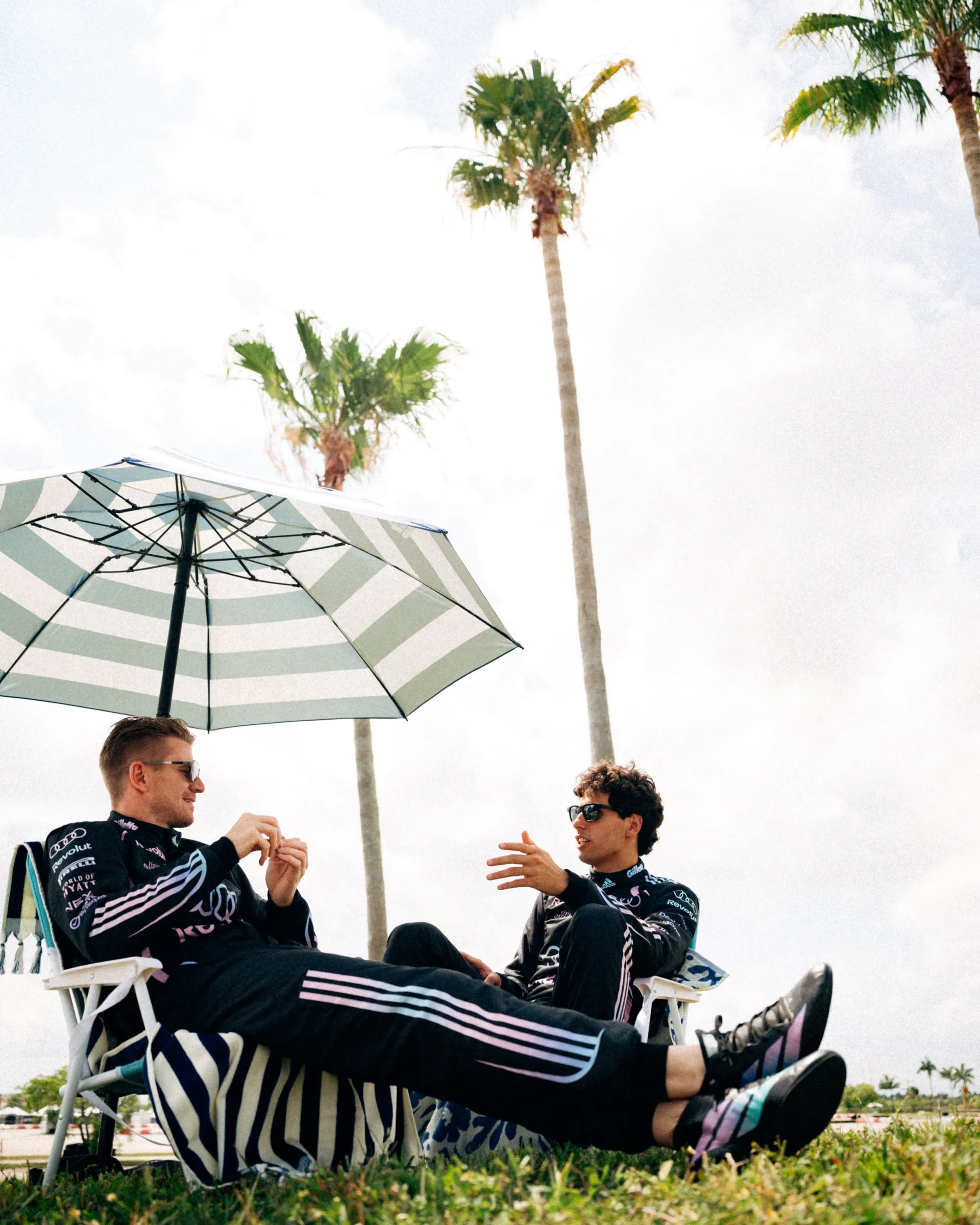 Nico Hulkenberg and Gabriel Bortoleto relax in beach chairs and talk beneath a striped umbrella, both wearing the Audi Revolut F1® Team Miami race suits on a grassy area lined with palm trees.
