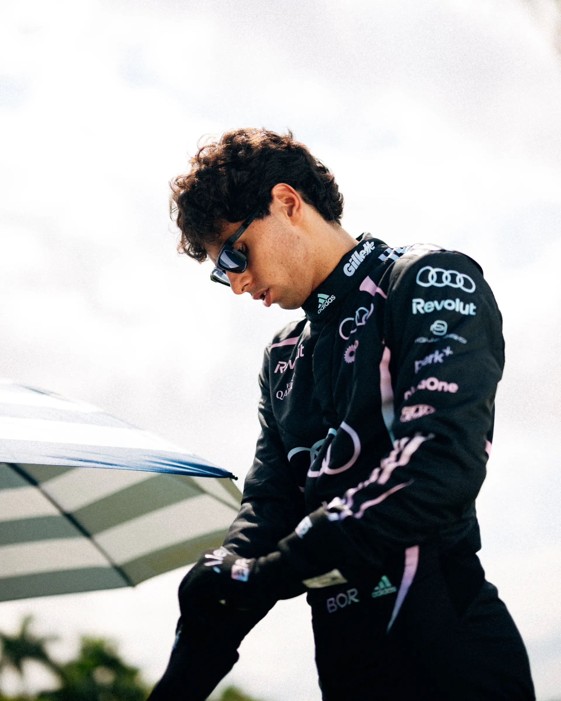 Gabriel Bortoleto looks downward while adjusting his gloves in the Audi Revolut F1® Team Miami race suit beside a striped beach umbrella.