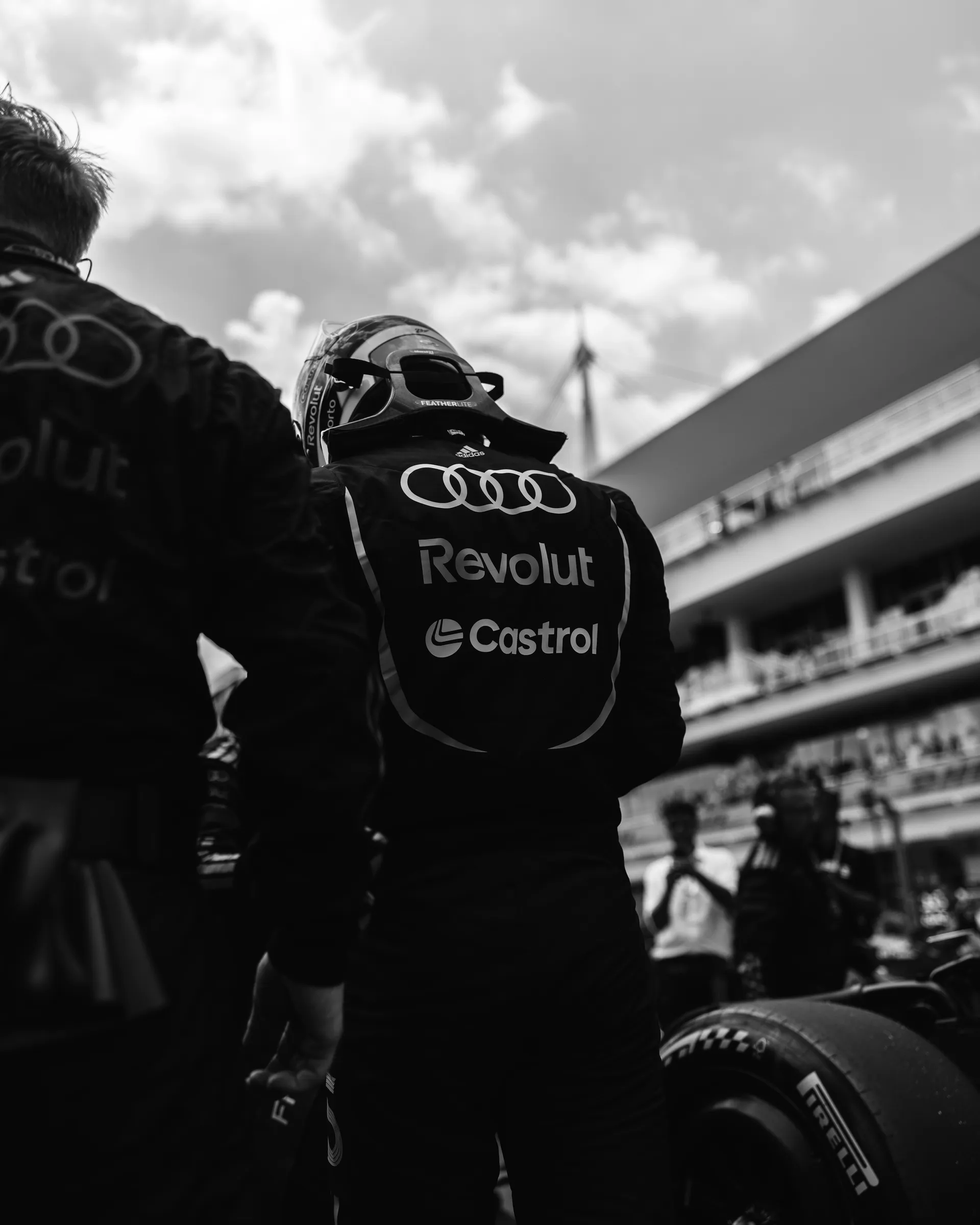 Black and white image of Gabriel Bortoleto standing beside the car from behind, wearing an Audi Revolut F1® Team race suit.