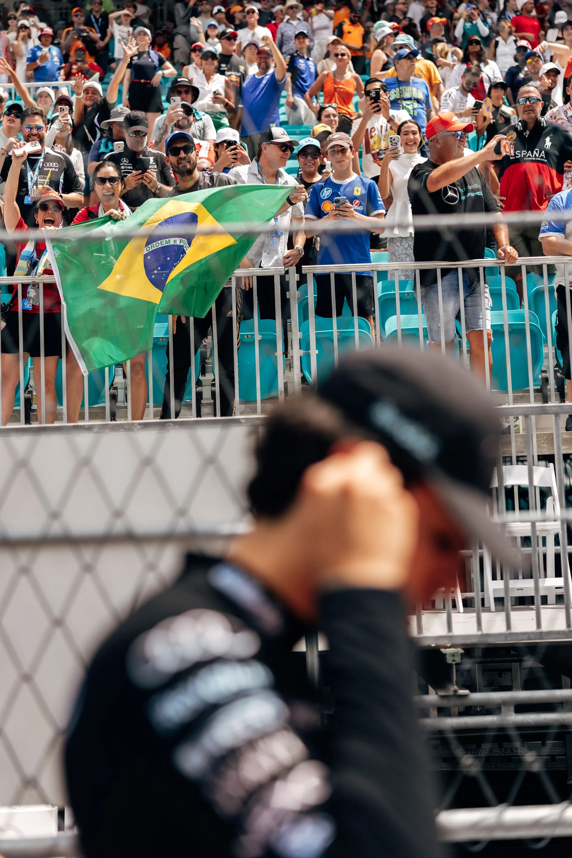Fans in the grandstand wave a Brazilian flag as a blurred Gabriel Bortoleto stands in the foreground.