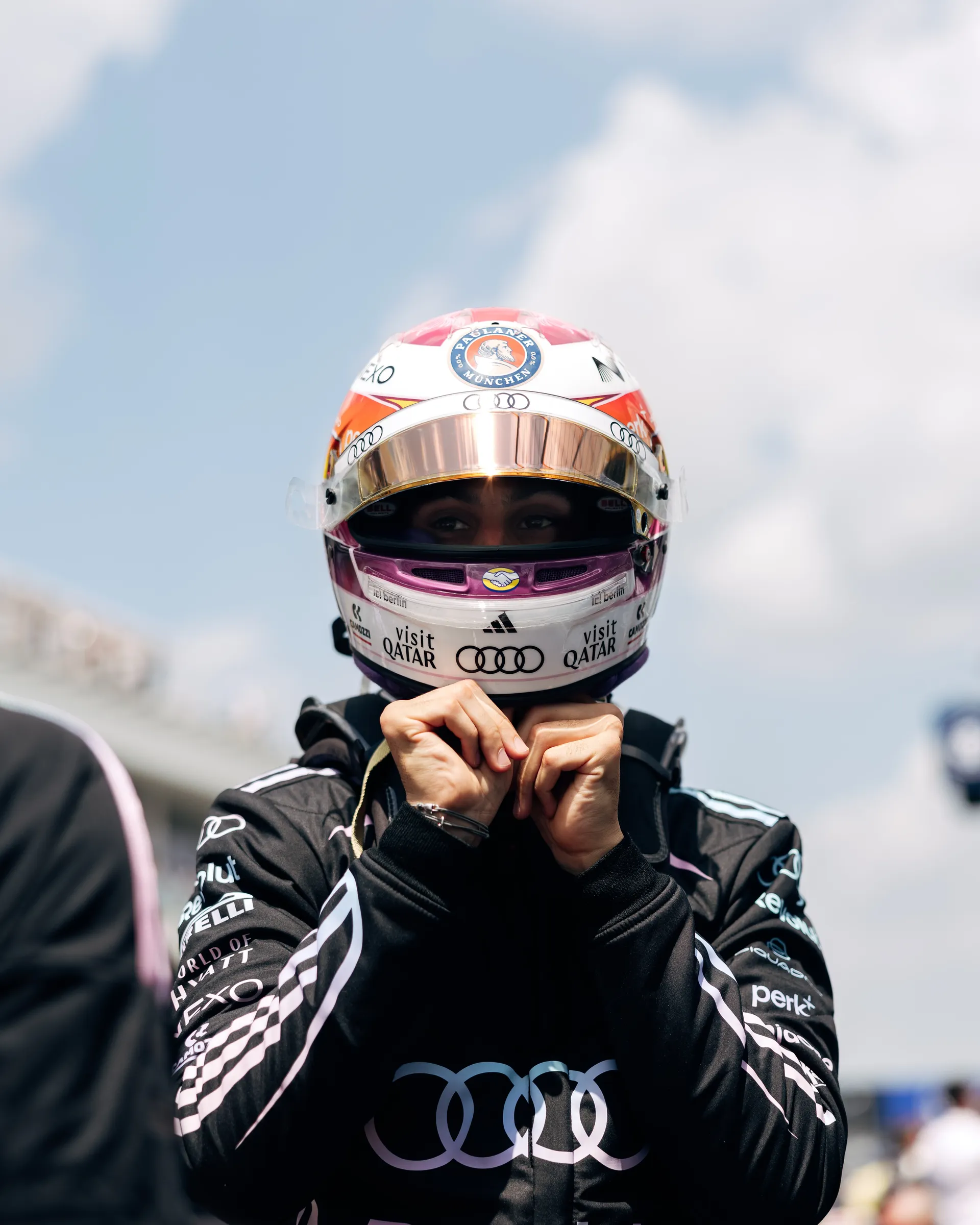 Gabriel Bortoleto faces the camera in his helmet and race suit while standing on the grid under a bright sky.