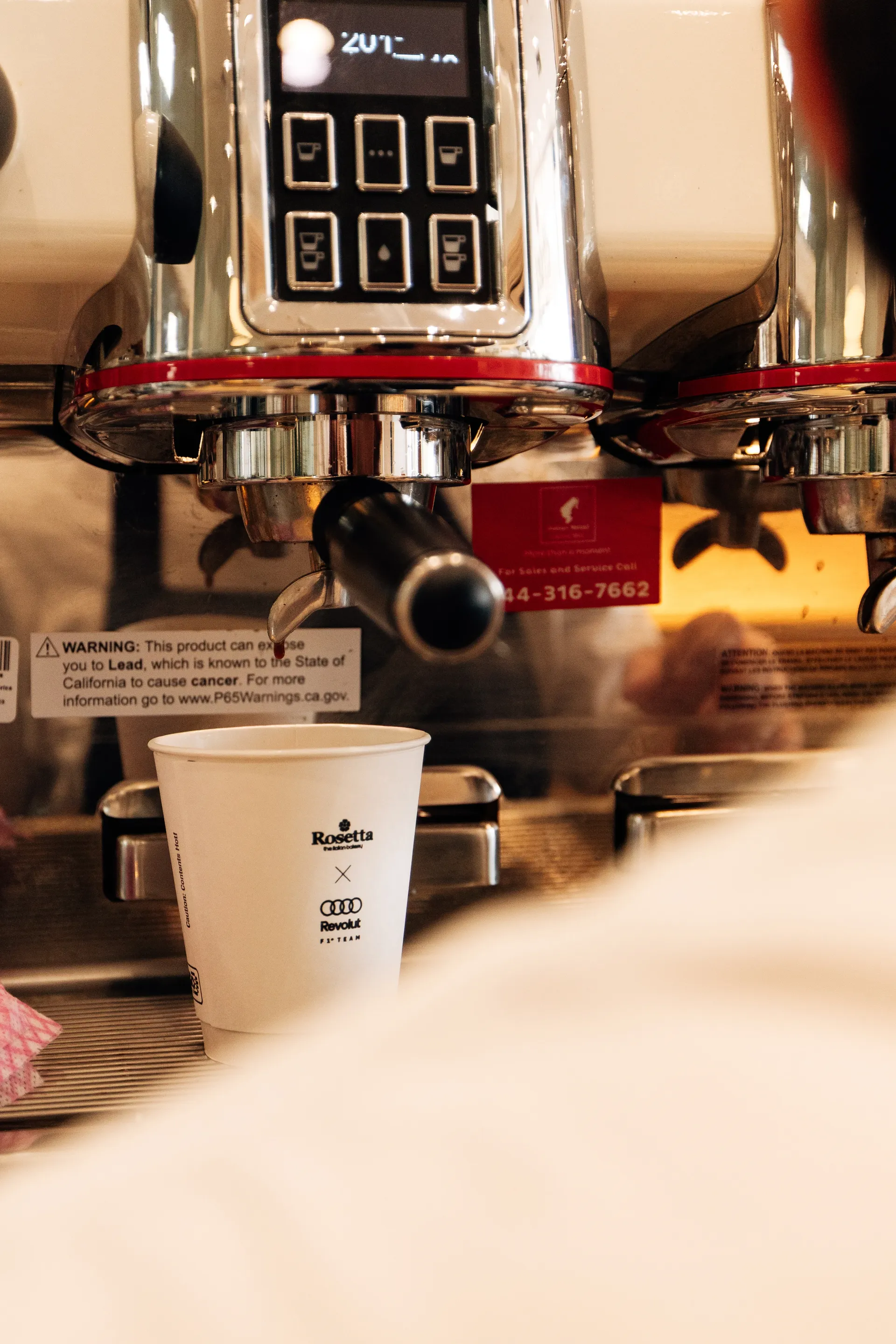 Close up of a coffee machine pouring into a white Rosetta x Audi Revolut F1® Team paper cup on the counter.