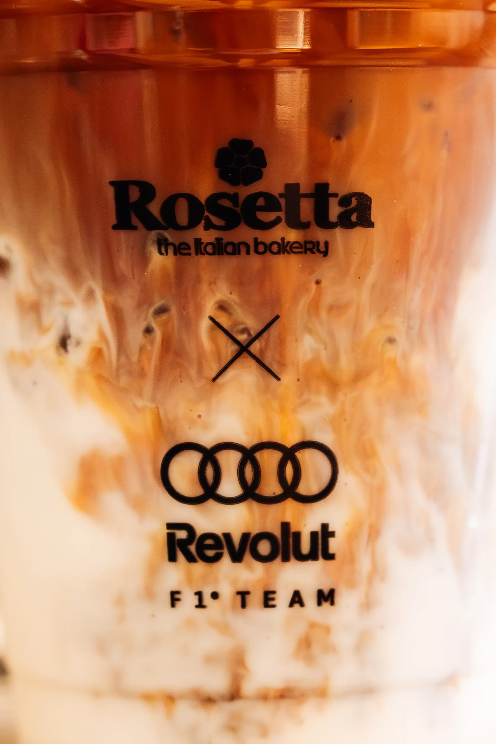 Close up of an iced coffee in a branded Rosetta x Audi Revolut F1® Team cup with cream swirling through the drink.