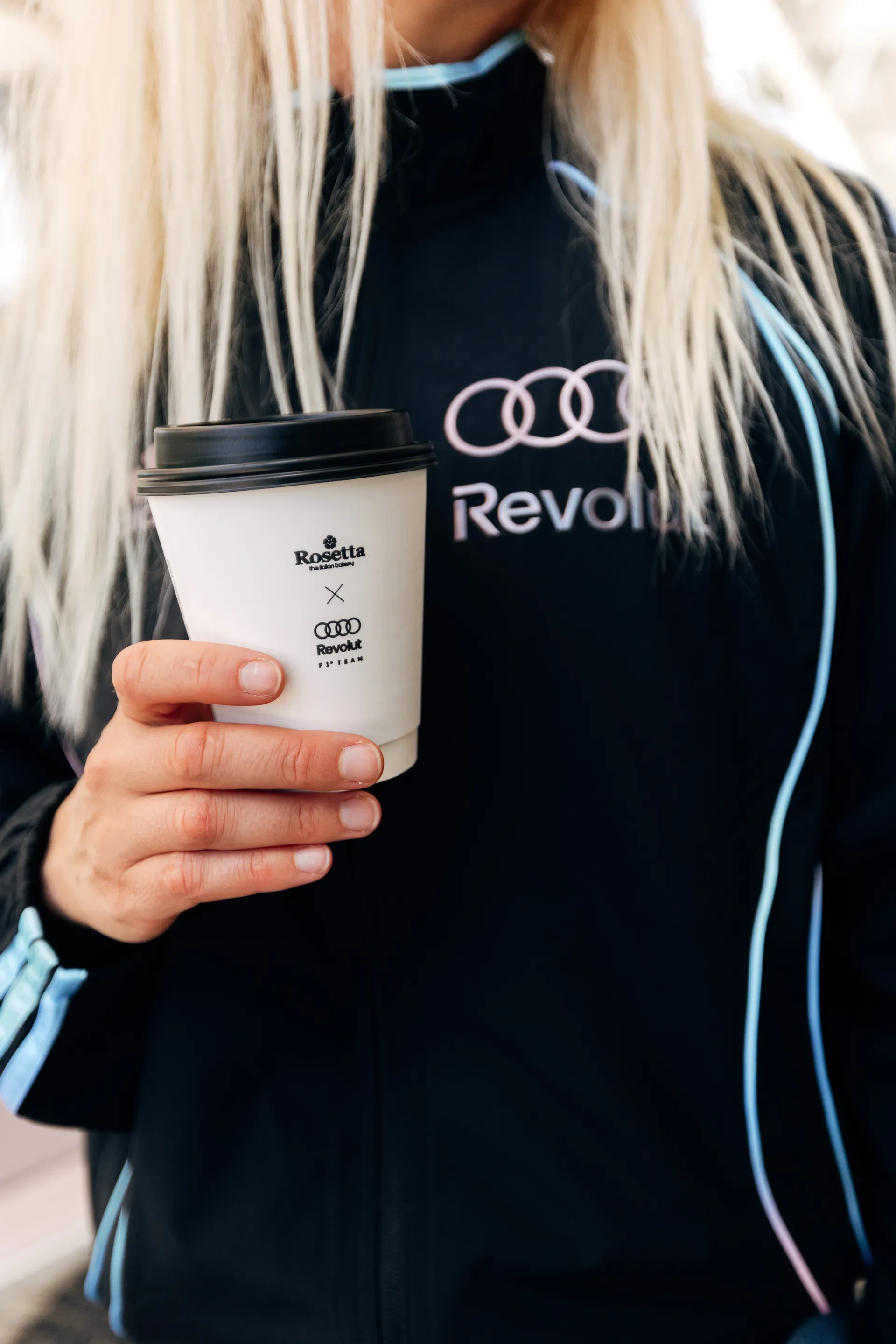 Person in a adidas x Audi Revolut F1® Team Miami collection track jacket holding a small branded takeaway coffee cup.