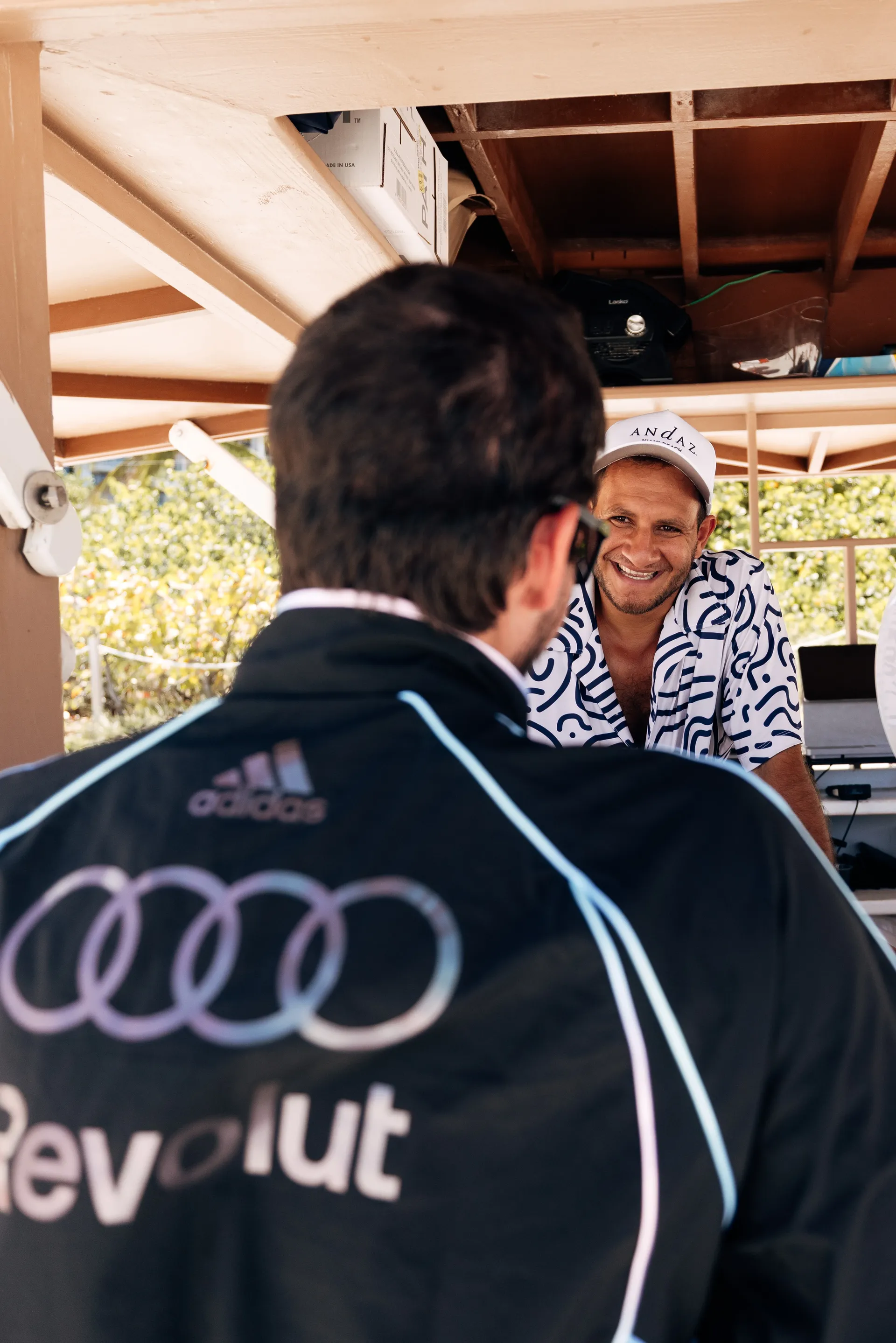 Person in a adidas x Audi Revolut F1® Team Miami collection jacket speaking with a smiling vendor at a beachside stand.