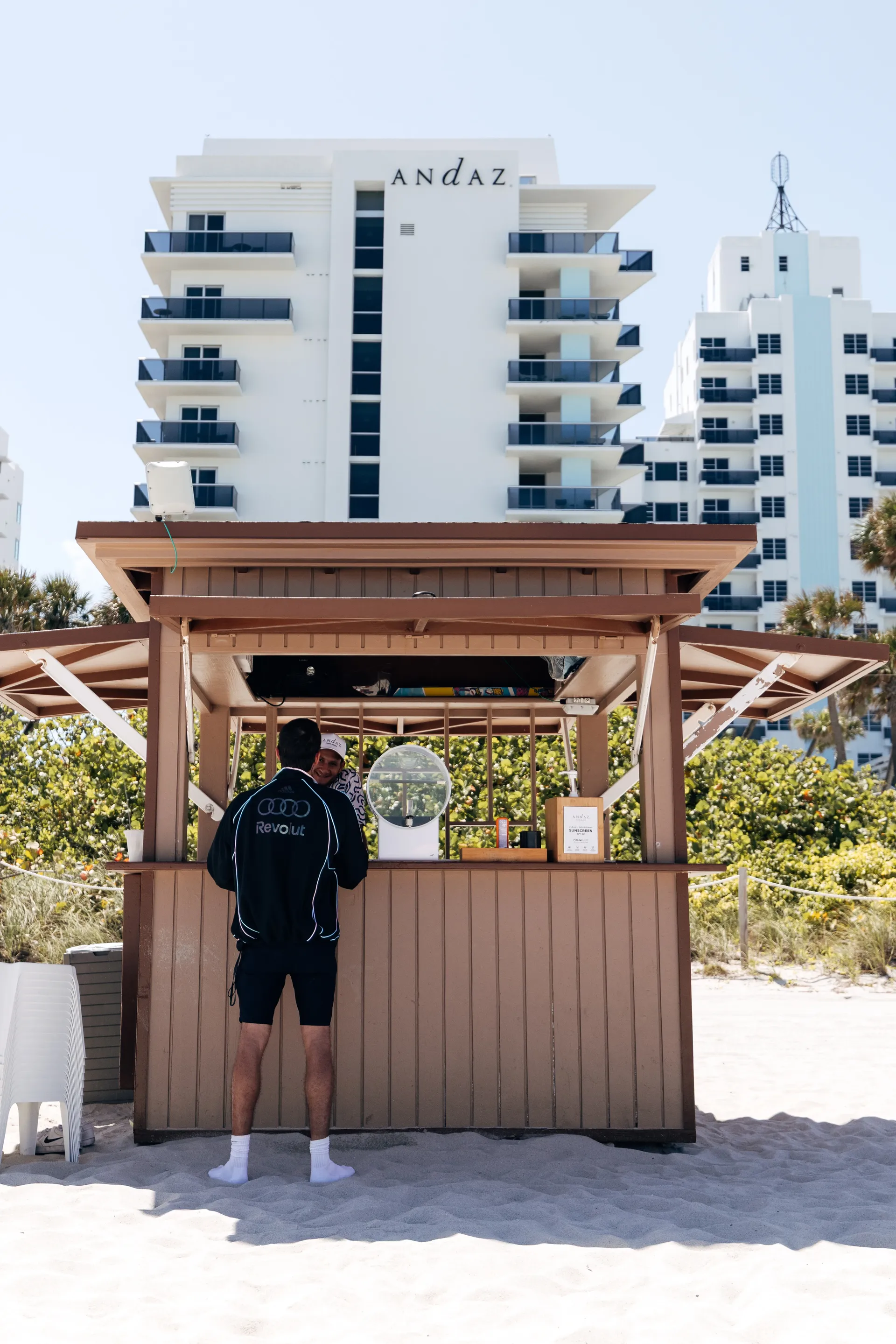 Wide shot of a beach kiosk with a person in a adidas x Audi Revolut F1® Team Miami collection outfit standing in the sand, with the Andaz hotel rising in the background.
