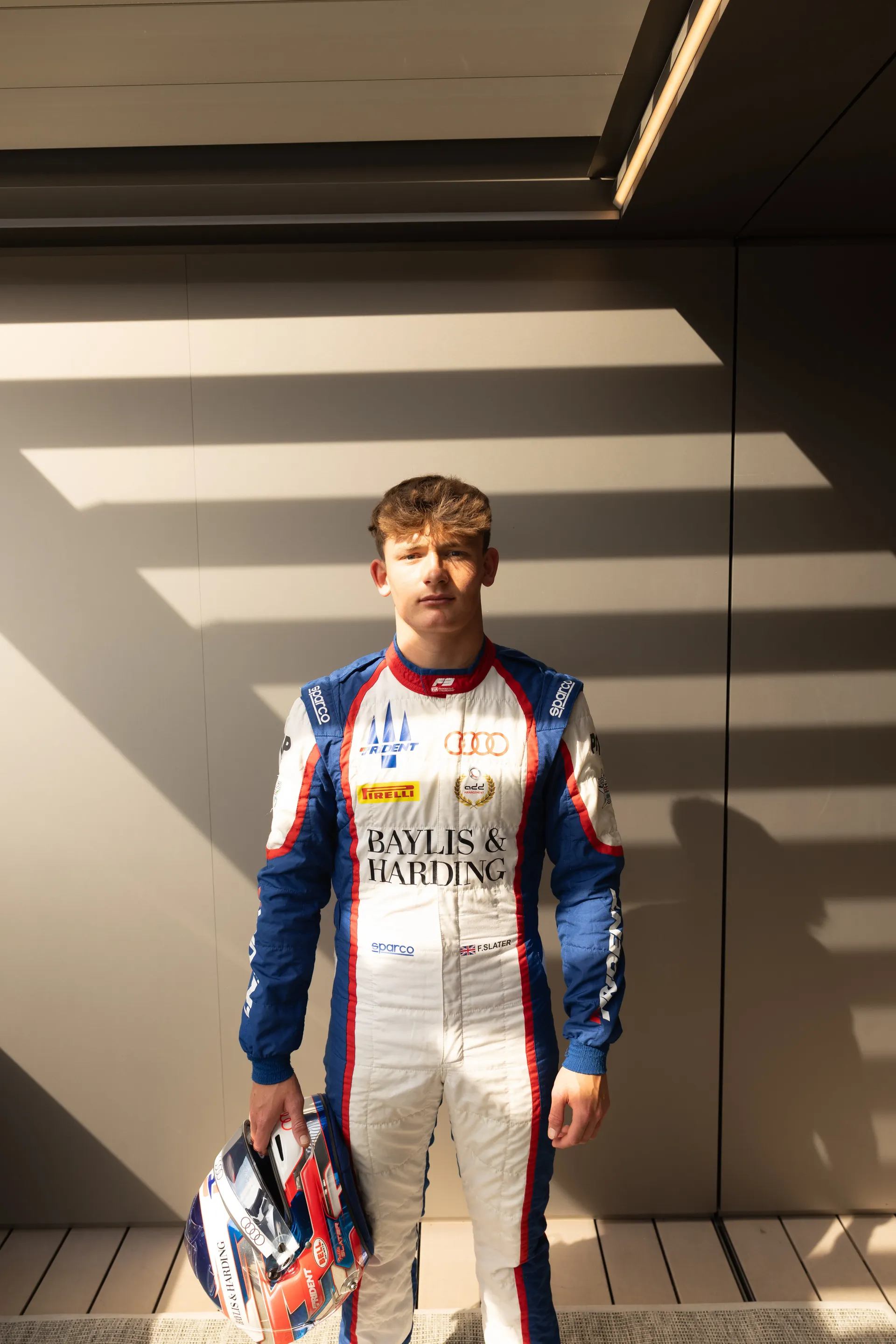 Freddie Slater in his race suit holding his helmet, striped light on the wall behind him