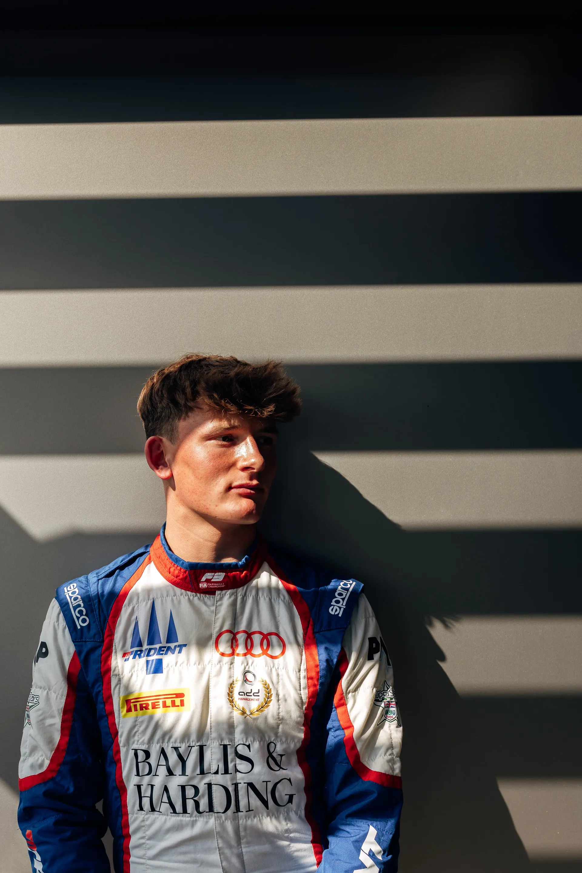 Freddie Slater in his race suit leaning against a wall, looking to the side