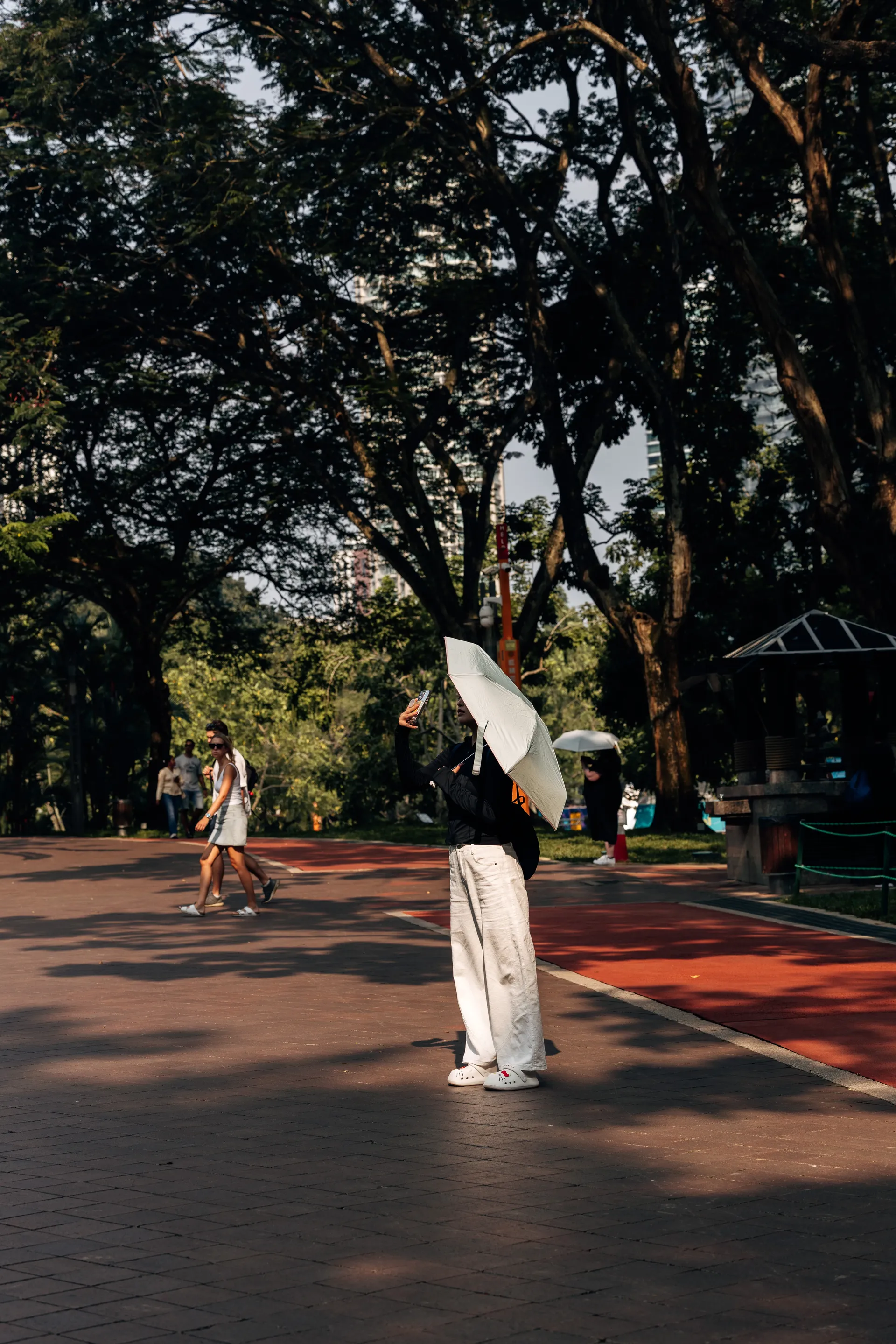 Park visitor with a white umbrella taking a photo on a paved path in Kuala Lumpur